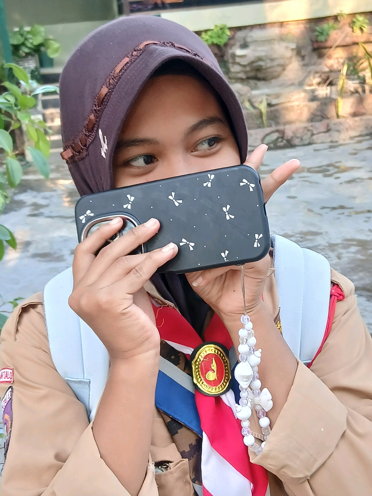 The image shows a young person wearing a brown scout uniform and a dark brown hijab. The person is holding a black smartphone with a white bow pattern in front of their face, obscuring most of their features. Their right eye is visible, looking to the right. Their right hand is holding the phone, and their left hand is extended with two fingers up in a peace sign. The person is wearing a red and white scarf, a blue sash, and a circular scout emblem with red and gold detailing. A white beaded lanyard with a charm is attached to the phone. The background appears to be outdoors, with stone steps and greenery visible. The overall lighting suggests it is daytime. The image does not provide any specific visual cues to pinpoint a location within Gorontalo, Indonesia.
