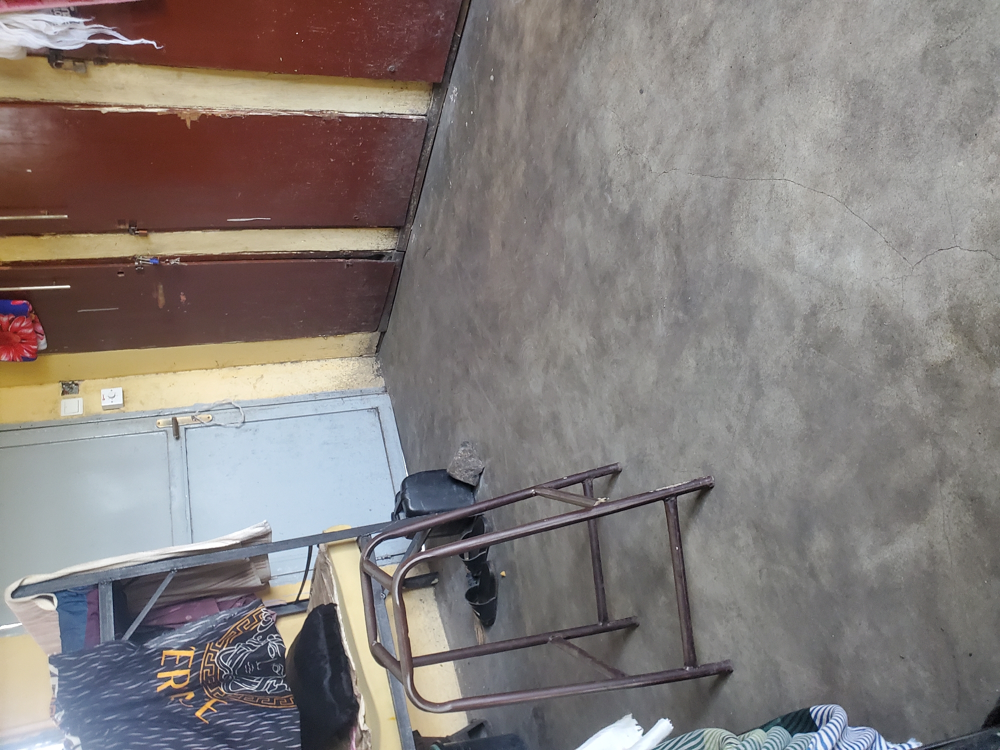 This image depicts an indoor scene, likely a room in Markurdi, Nigeria. The room appears utilitarian and somewhat cluttered. Dominating the left side of the frame are two sets of dark brown, wooden double doors, each with visible hardware and a yellow-painted trim. One set of doors is slightly ajar, revealing a glimpse of what might be a metallic surface behind it. To the right of the doors, a light grey metal door with a handle is visible. Above this door are two light-colored electrical fixtures, one resembling a light switch.
In the foreground and middle ground, a metal frame structure, possibly a cot or shelving unit, is present. It is made of brown metal tubing and appears somewhat aged. On this structure, several items are placed, including folded fabrics and what seems to be a mattress or bedding. A black garment, possibly trousers, is also visible. To the right of this structure, a small, dark seat with a stone placed on top of it is visible. A pair of black shoes are positioned near the seat.
The floor is a concrete surface, showing signs of wear and a few cracks. The overall lighting suggests an indoor environment, possibly during the daytime, though no direct light source is evident. The scene conveys a sense of everyday living in a possibly modest setting. There is no clear indication of any specific activity taking place, and the overall impression is one of stillness. The text "ER" is partially visible on a patterned fabric in the foreground.