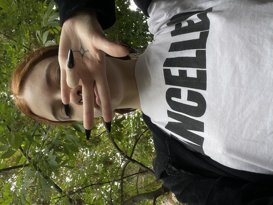 A young individual, appearing to be female with reddish-brown hair, is captured in a close-up outdoor photograph from a low angle. She wears a white t-shirt with large black block letters, partially obscured, which spell "ANCELLE," likely part of the word "CANCELLED." Over the t-shirt, a black, unzipped fleece jacket or hoodie is worn. Her left hand is raised, with fingers spread, partially covering her face, specifically obscuring her eyes and nose bridge. The index and middle fingers are extended, appearing to cover her eyes, while the ring finger and pinky are curled. She has long, sharply pointed black artificial fingernails on all visible fingers. A small, black star-like tattoo is visible on the palm side of her left hand, near the base of her middle finger. A silver septum piercing is visible in her nose. The background consists of dense green foliage, indicating trees or shrubs, suggesting a park or heavily treed area. The lighting appears soft and diffused. The specific location within the city of Buenos Aires, Argentina, cannot be determined from the visual information provided in the image.