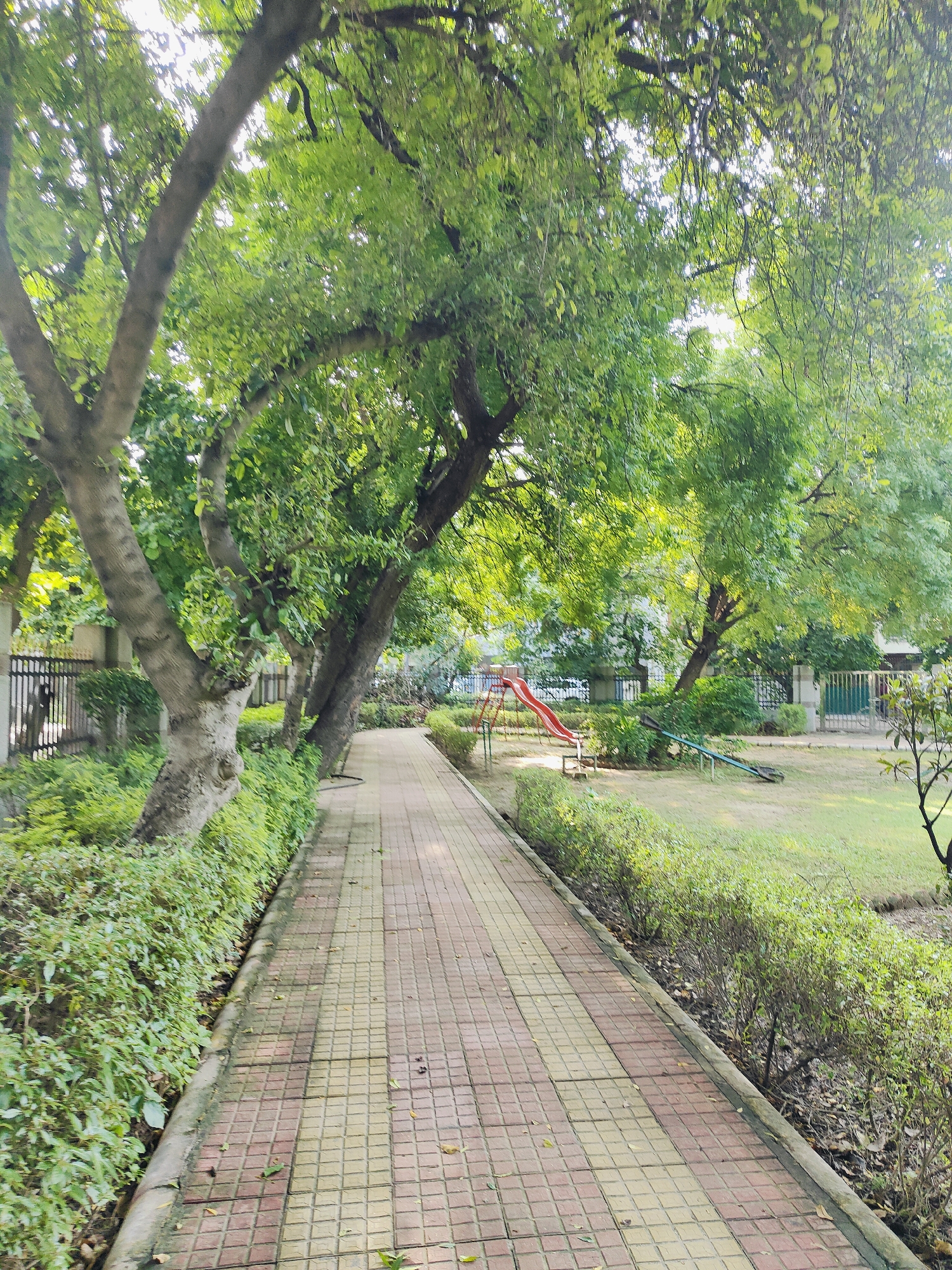 The image depicts a pathway in a park in Gurgaon, India. The pathway is paved with red and yellow square tiles and is bordered by lush green bushes and trees. Sunlight filters through the dense foliage of the trees, creating dappled shadows on the path. In the background, playground equipment, including a red slide and a seesaw, is visible on a grassy area. The park appears to be well-maintained, with a fence lining the perimeter in some sections. No people are present in the scene. The overall impression is one of a tranquil, urban green space.