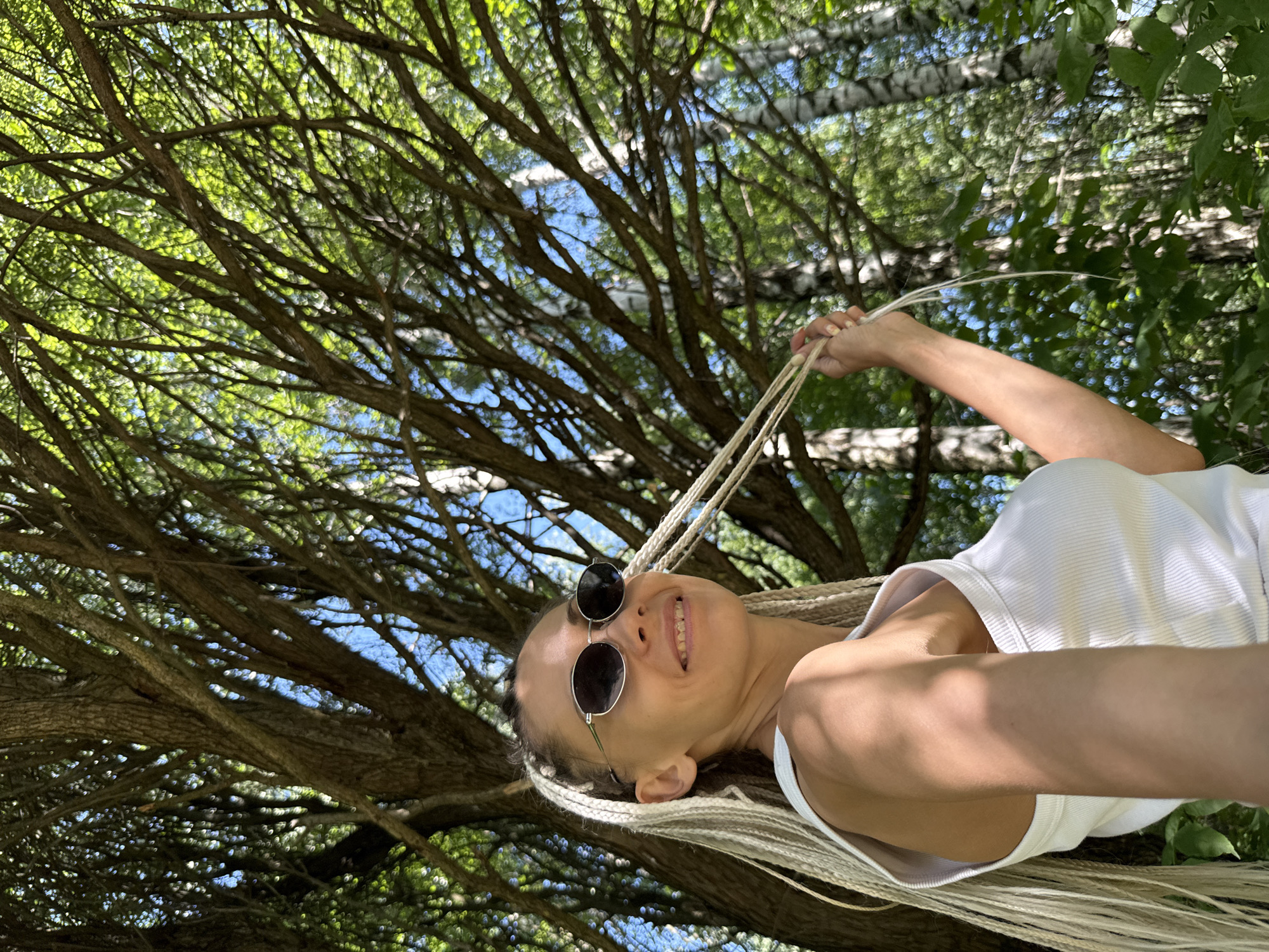 A woman with blonde braided hair and sunglasses is standing outside in a wooded area. She is wearing a white tank top and holding a rope in her right hand. The trees in the background are tall and have green leaves. The lighting suggests it is daytime and sunny. The scene appears to be in Rusanovo, Russia, based on the provided information.