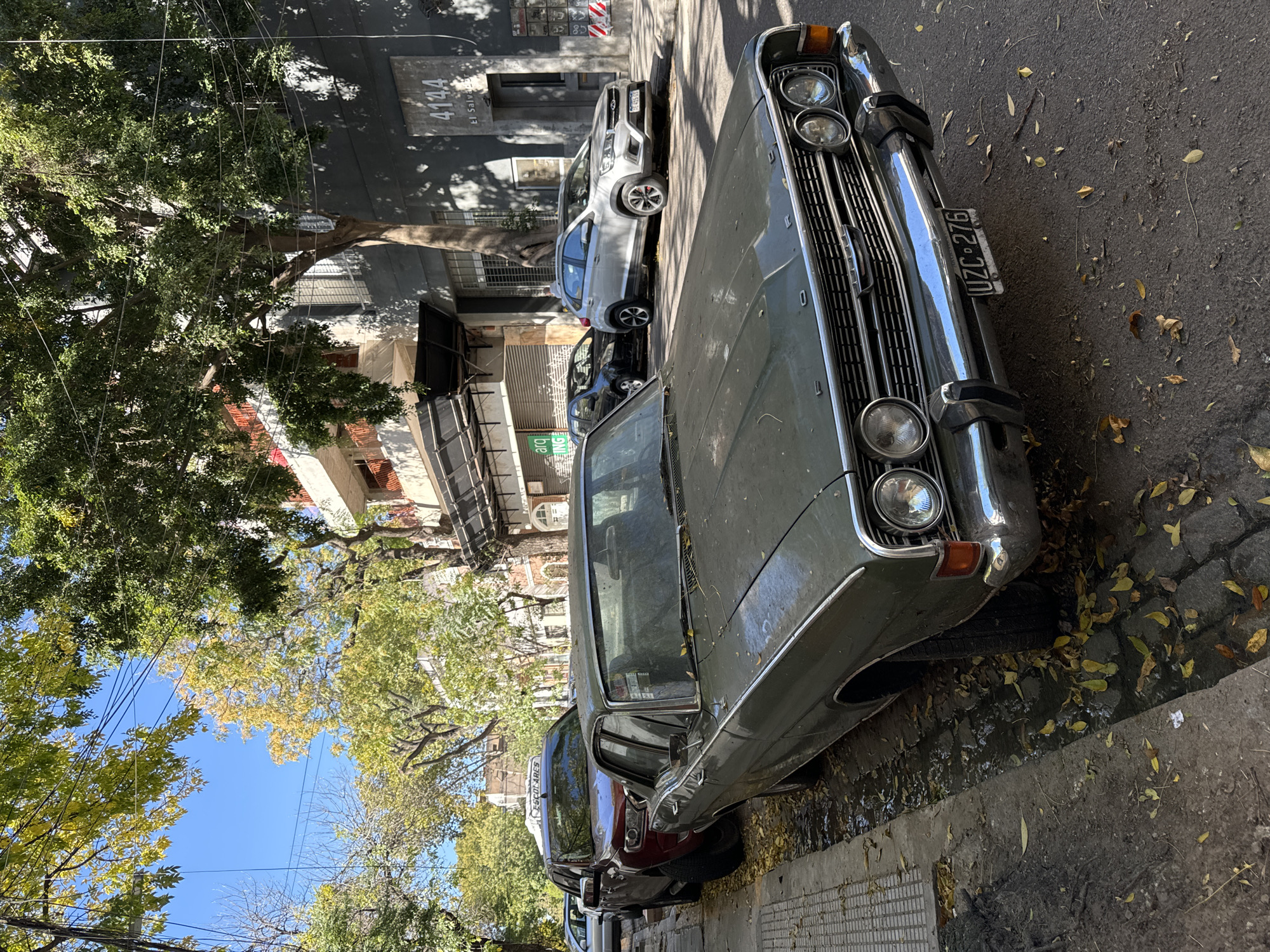 The image depicts a bright, sunny street scene in Buenos Aires, Argentina, likely during autumn given the fallen leaves. The primary subject is an older, dark green sedan, possibly a classic car, parked on the right side of the street, facing forward. It features prominent chrome bumpers, a large grille, and four round headlights. Its license plate, "UZC-276", is clearly visible on the front bumper.
Behind the green sedan, a dark red car is parked, followed by a modern, light-colored SUV, whose license plate partially shows "AE053". Other vehicles are parked further down the street. The street itself is lined with trees displaying a mix of green and yellowish autumnal foliage, casting strong shadows across the road and buildings. A clear blue sky is visible through the tree canopy, crisscrossed by overhead wires.
Buildings line the street, with one facade in the background prominently displaying the number "4144" and the street name "El Salvador", confirming the location. The asphalt road and concrete sidewalk are covered with scattered yellow and brown leaves, and some dirt and damp patches are visible near the curb. No people are present in the scene. A small green sign with the text "arq IMG" is visible on a building. The time of day appears to be mid-morning or early afternoon due to the bright sunlight and shadow angles.
