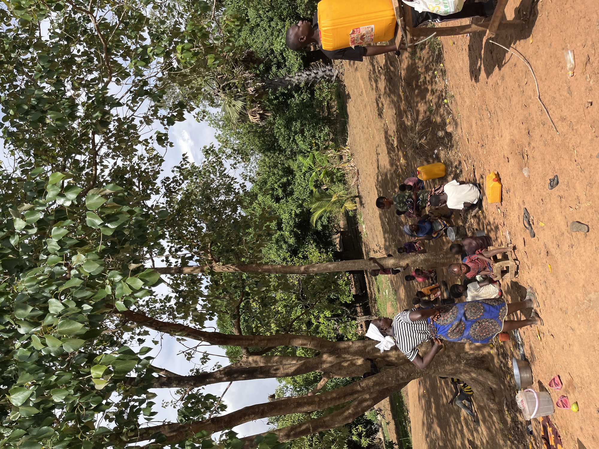 A group of people, including children, gather in a dirt clearing beneath trees in what appears to be Barakin Momo Dogo, Nigeria. Several yellow jerrycans sit on the ground near the group. One man stands near a table, holding a yellow jerrycan aloft. A woman in a patterned dress stands by a tree with a bucket nearby. The scene is bathed in sunlight with shadows cast by the trees. The ground is dirt and sandy in appearance.