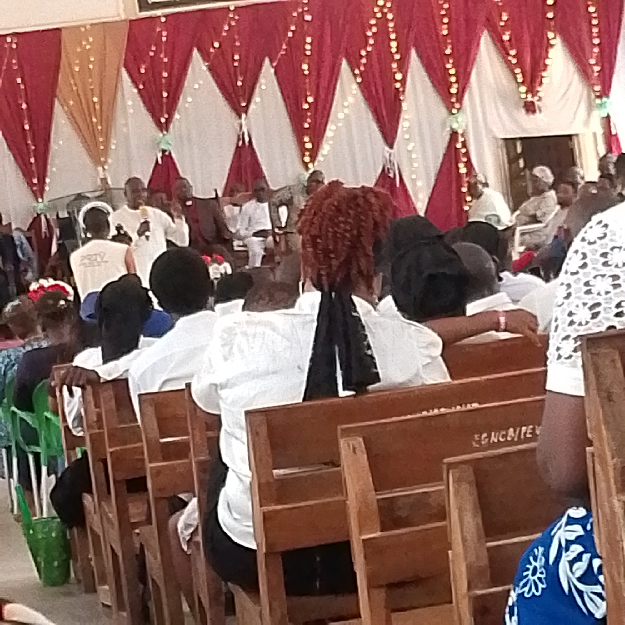 The image depicts an indoor gathering of people seated in wooden pews facing a stage-like area. In the front, individuals are seen from the back, mostly wearing white shirts. In the foreground, brown pews are visible, some with markings or labels. Further away, on a slightly elevated area, are several men, one of whom is speaking into a microphone. Behind them is a backdrop of draped fabric in shades of red, white, and gold, adorned with strings of lights. The location appears to be a church or similar gathering space.