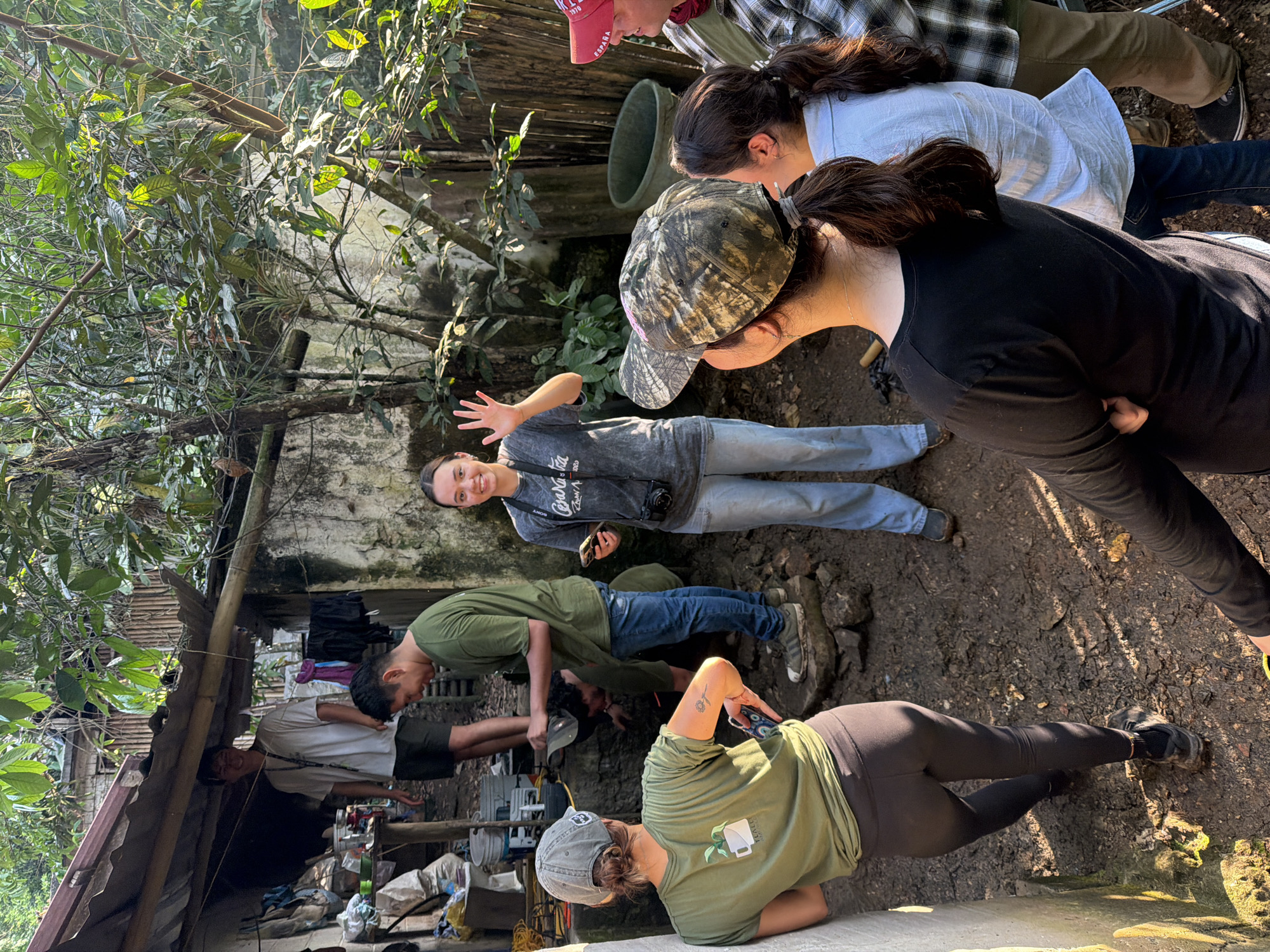 A group of people are gathered outdoors in Tetelilla de Islas, Mexico. Some individuals are looking down at something on the ground while others are standing. One woman is smiling and waving towards the camera. There is a building visible in the background with a corrugated metal roof and clothes hanging outside. The surrounding environment includes vegetation and dirt.