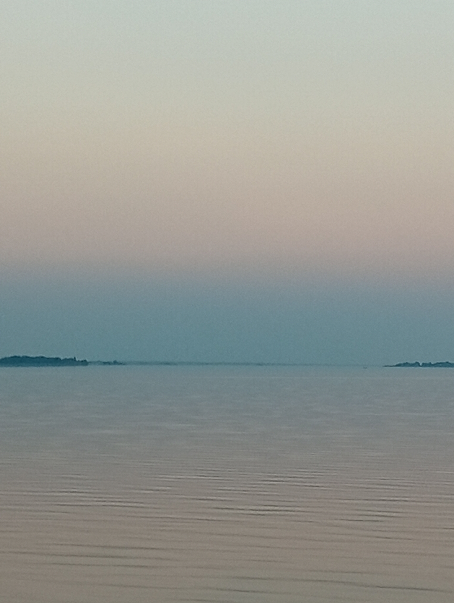 The image depicts a serene, expansive body of water under a gradient sky transitioning from a soft pinkish-orange at the top to a pale blue near the horizon. The water's surface is calm, with subtle horizontal ripples creating a textured effect. On the distant horizon, two small, dark landmasses are visible, one on the left and another on the right, appearing as islands or peninsulas. The overall atmosphere is peaceful and tranquil, suggesting a sunrise or sunset. The location is identified as Unknown, Unknown.