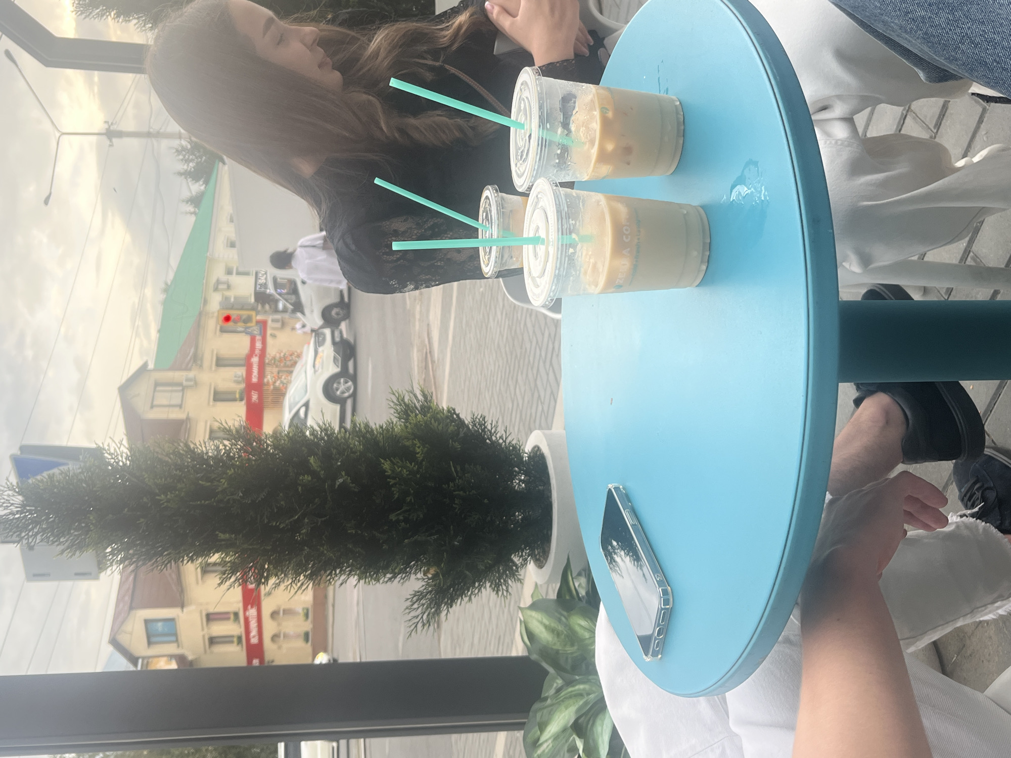 The image depicts an outdoor seating area at a cafe in Oskemen, Kazakhstan, with a bright turquoise table in the foreground. On the table, there are three clear plastic cups with iced beverages and light-colored straws, two of which are in the foreground and one slightly further back. A smartphone in a clear case is placed on the table. To the right, a young woman with long, dark, wavy hair, wearing a black lace top, is partially visible, looking down. In the background, beyond the cafe's seating area, a street scene is visible with buildings, cars, and pedestrians. A tall, conical green shrub is positioned behind the table. The sky is overcast.
