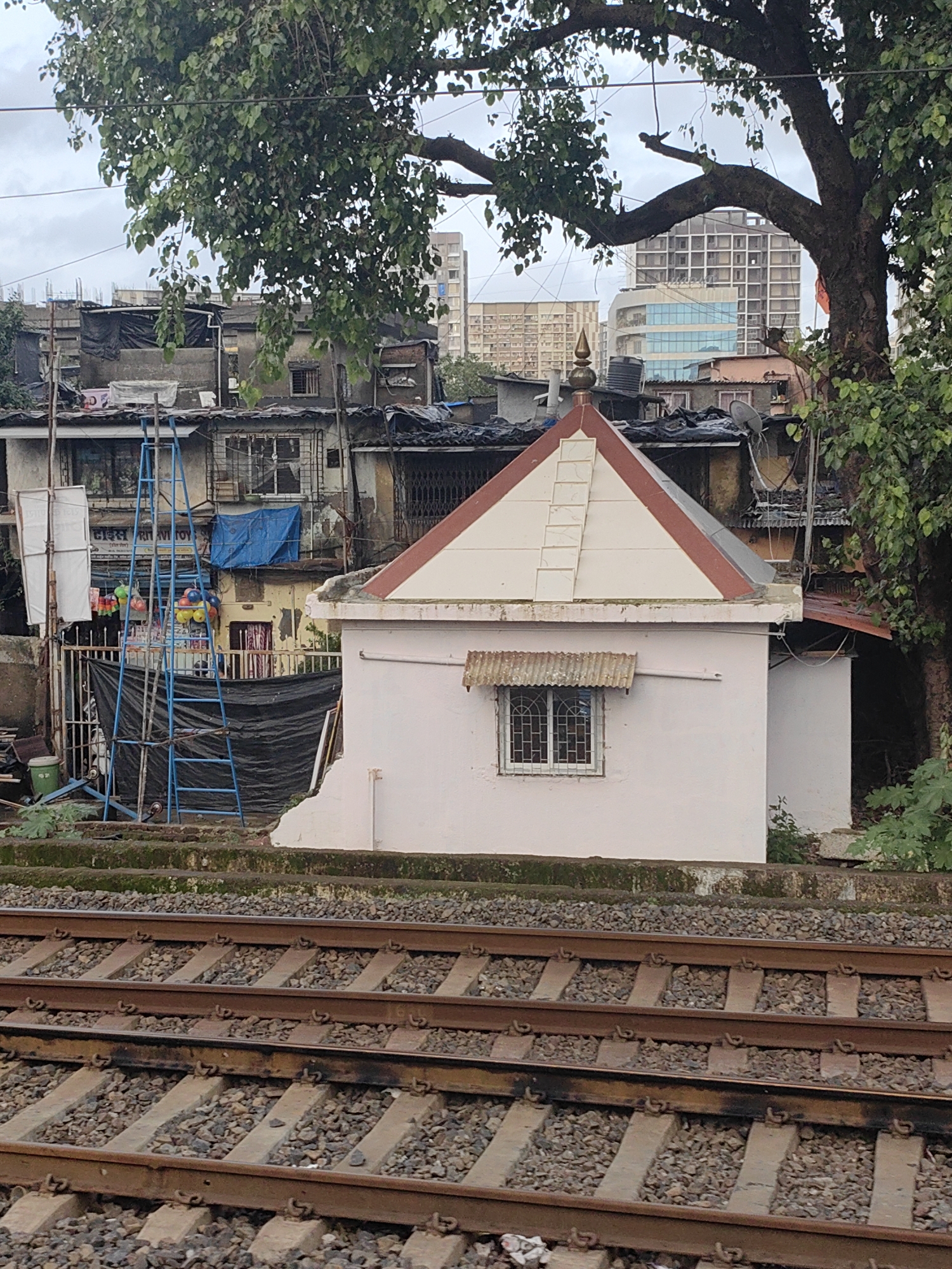 The image depicts a view of railway tracks in Mumbai, India, with a small building resembling a temple in the foreground. The building has a white facade with a triangular red-and-white roof, topped with a golden spire. A small, barred window with a corrugated metal awning is visible on the front of the building. Behind the temple, a collection of tightly packed, multi-story residential buildings are visible, characteristic of a densely populated urban environment. These structures appear weathered and some have tarpaulins or corrugated metal sheeting as roofing. In the mid-ground, a blue ladder stands against one of the residential buildings, and some colorful balloons are suspended from it. A large tree with dense foliage overhangs the scene, casting shadows. The railway tracks in the foreground are made of concrete sleepers and ballast. In the background, modern high-rise buildings can be seen, suggesting a mix of old and new development within the city. The sky is overcast.