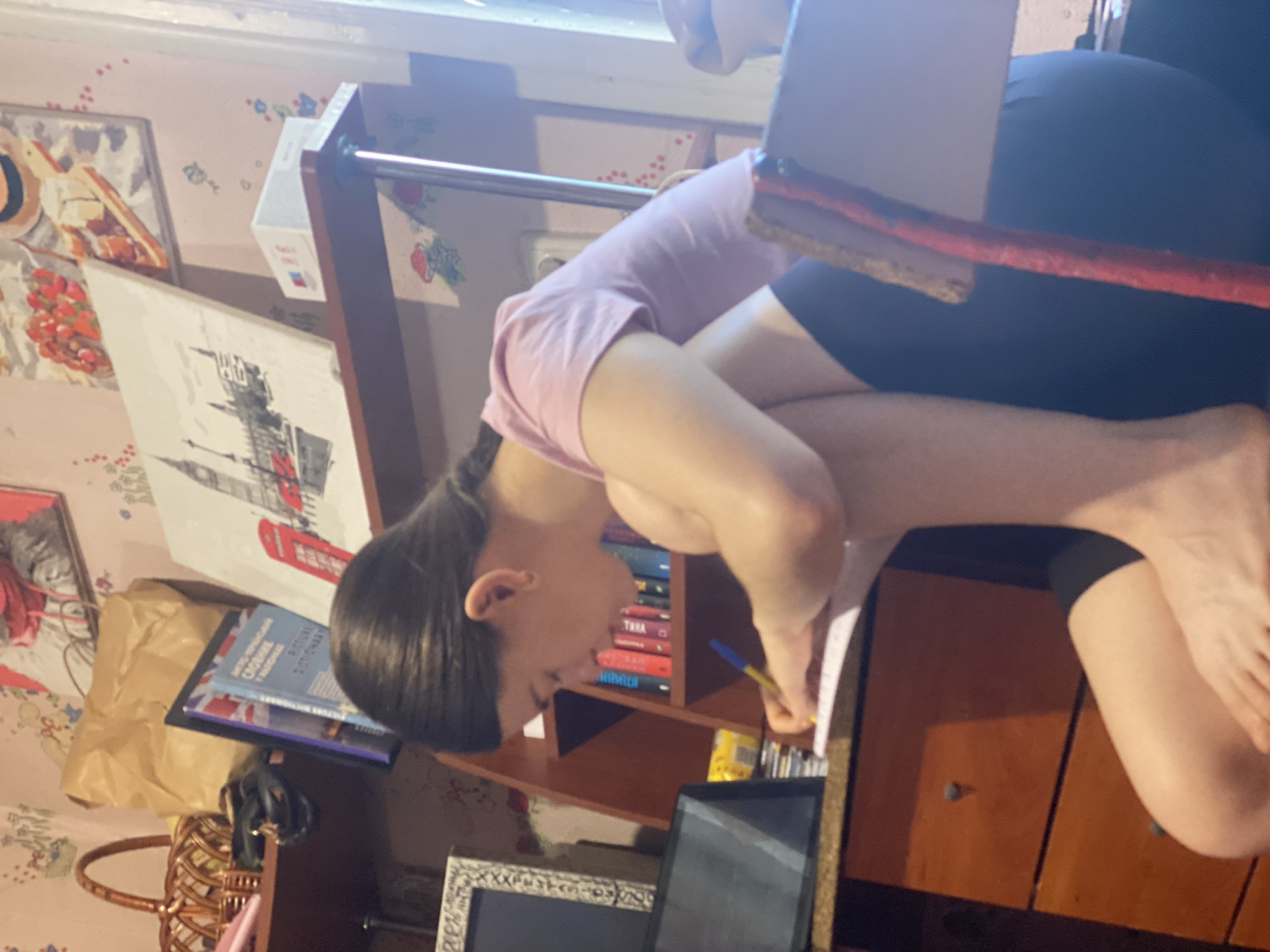 A young person with dark hair, wearing a pink t-shirt and black shorts, is sitting at a desk and writing in a notebook. They are hunched over the notebook, with their right hand holding a pen. A tablet is visible on the desk to the left of the notebook. In the background, there is a shelf filled with books and decorative items, including a painting of a red telephone box and Big Ben. The room appears to be a study or bedroom, with patterned wallpaper and wooden furniture. The lighting suggests it is daytime, with light coming from a window on the right side of the image.