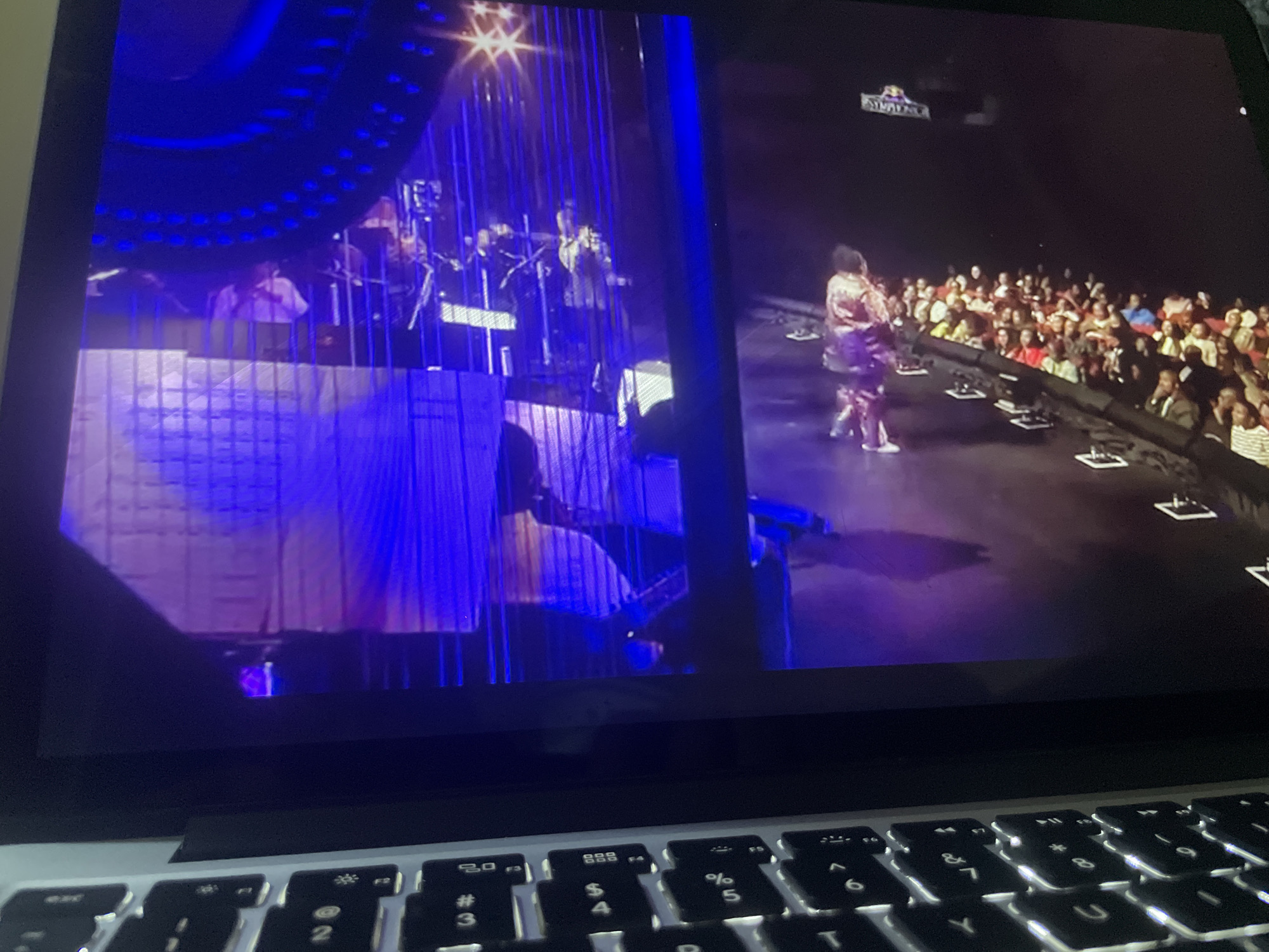 The image displays a laptop screen showing a live performance. On the left side of the screen, a band is visible on a stage with blue lighting and a checkered backdrop. In the center of the stage, a person in a white shirt appears to be singing. On the right side of the screen, a performer in a shimmering outfit is dancing in front of a large audience. The audience is seated and facing the stage, with some individuals appearing to clap. The overall scene suggests an indoor concert or performance event. The location in the city of Howayi, Nigeria, is not identifiable from the image content.