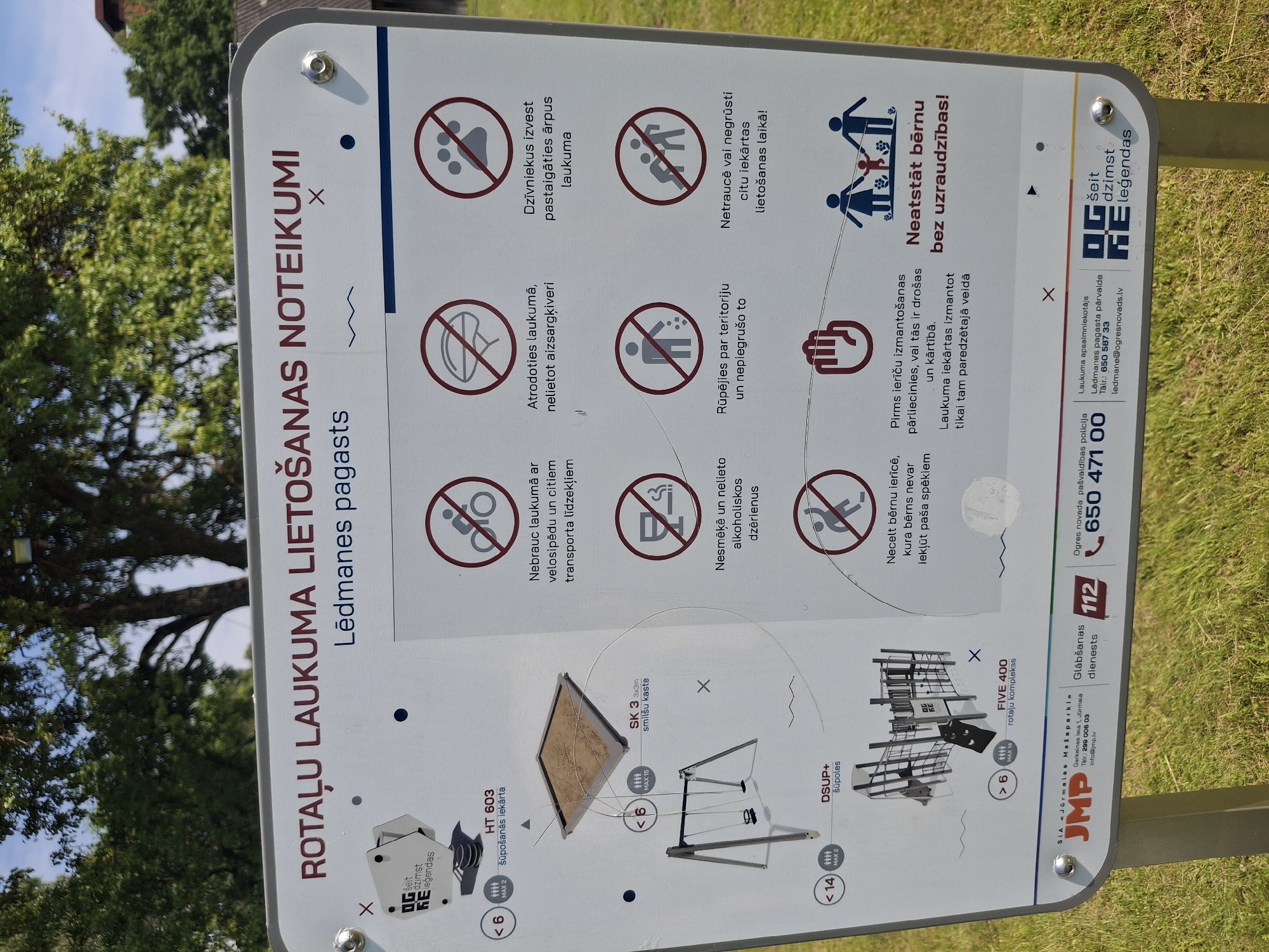 A white sign with red text and icons is mounted on a grassy area. The sign is titled "ROTĀLU LAUKUMA LIETOŠANAS NOTEIKUMI" which translates to "PLAYGROUND USAGE RULES." Below this, it specifies "Lēdmanes pagasts," indicating the location is Lēdmane parish.
The sign features various pictograms illustrating rules for playground use. These include:
- A bicycle with a line through it, indicating no bicycles.
- A helmet with a line through it, suggesting the use of protective gear.
- A paw print with a line through it, prohibiting animals.
- A cigarette and drink with lines through them, meaning no smoking or alcoholic beverages.
- A person littering with a line through it, emphasizing keeping the area clean.
- Two people with a line through them, possibly indicating no roughhousing or aggressive behavior.
- A child with a magnifying glass and a line through it, possibly meaning children should be supervised.
- A family group with a "Neatstāt bērnu bez uzraudzības!" caption, which translates to "Do not leave children unattended!"
On the left side of the sign, there are diagrams of playground equipment with age and maximum user restrictions. For example, there's a sandpit labeled "SK 3 3x3m smilšu kaste" (SK 3 3x3m sandbox) with a restriction of "< 6" and "MAX 15," and a swing set labeled "DSUP+ šūpoles" (DSUP+ swings) with "< 14" and "MAX 2." There is also a larger play structure labeled "FIVE 400 rotālu komplekss" (FIVE 400 playground complex) with "> 6" and "MAX 14."
At the bottom of the sign, there are logos and contact information for local services. This includes "SIA Jūrmalas Mežaparki" (Jūrmala Parks LLC), a phone number, an email address, "Glābšanas dienests 112" (Rescue Service 112), and information for "Ogres novada pašvaldība" (Ogre Municipality).
The background of the image shows green grass and trees, with a blue sky overhead. The sign appears to be in good condition, with some slight wear.