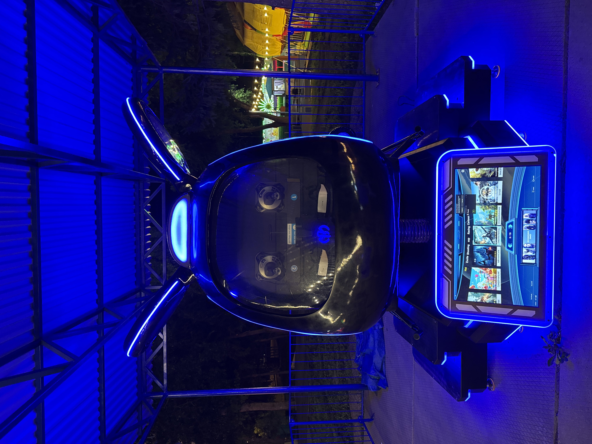 This is a medium shot, eye-level, outdoor, nighttime photograph of a futuristic-looking ride at an amusement park in Oskemen, Kazakhstan. The ride's main structure is a large, dark, egg-shaped pod with two wing-like appendages extending from the top, both lined with blue LED lights. The pod has a black, reflective surface, and two large circular lenses on the front suggest it is a simulator or virtual reality attraction. Below the pod is a base with more blue LED lighting. A touchscreen interface is mounted at the front of the base, displaying game tiles and menus. The ride is situated on a patterned ground surface under a covered structure with a corrugated blue roof supported by metal beams. Beyond the ride, a fence separates it from a background of trees and an amusement park with colorful lights and structures, including what appears to be a Ferris wheel and a yellow tent. The overall impression is one of a modern, technologically advanced amusement ride illuminated by striking blue lights in a park setting at night.