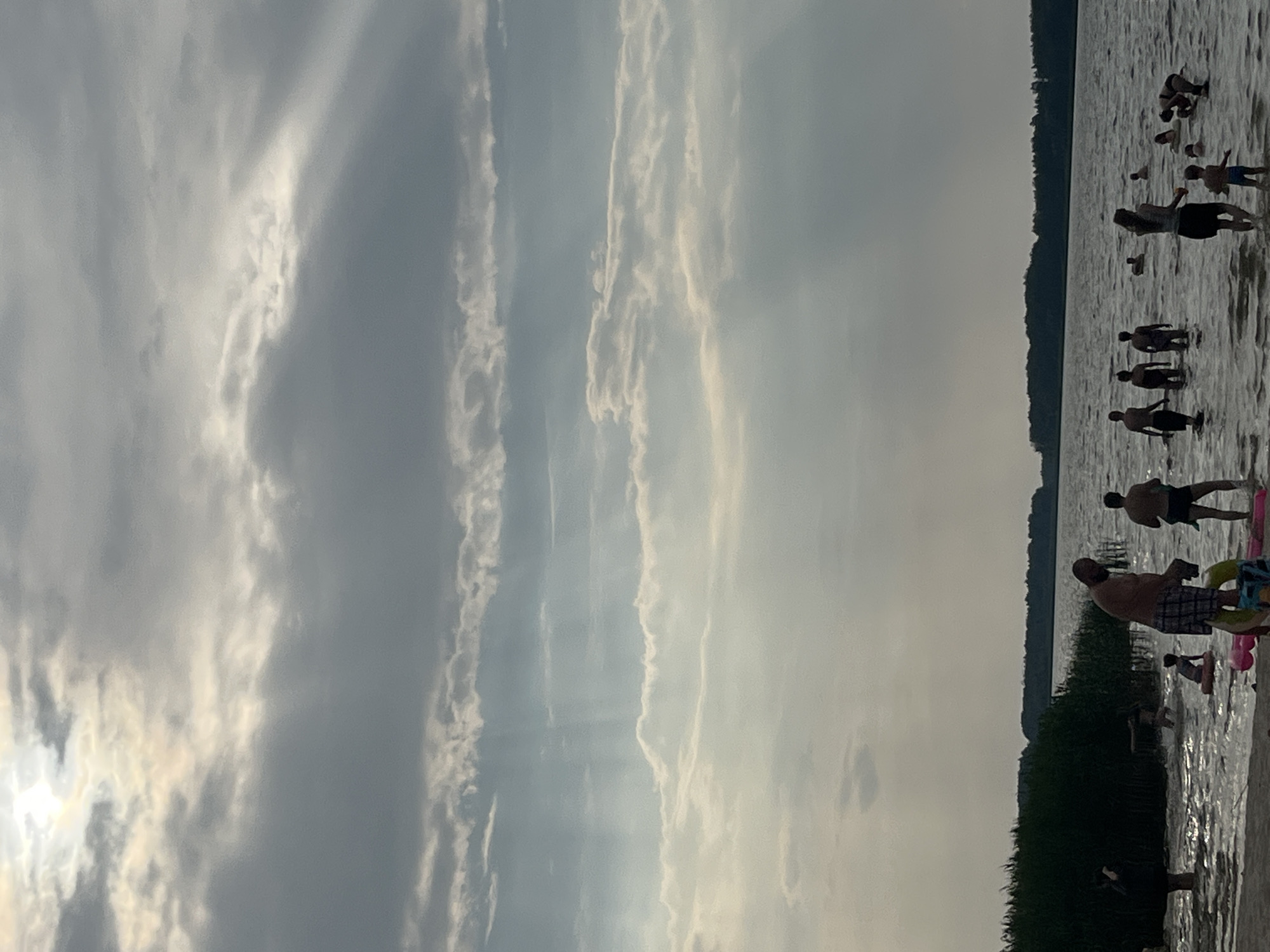 The image depicts a serene summer day at a lake in Загародскі сельскі Савет, Belarus. The sky is filled with dramatic, sun-dappled clouds, with rays of light piercing through, creating an ethereal atmosphere. In the foreground, a group of people are enjoying the water. Several individuals, including adults and children, are wading and swimming in the lake. Some are standing in the shallow water near the sandy shore, while others are further out. There are inflatable toys visible in the water. The shoreline is lined with dense green reeds and trees, forming a natural backdrop. The lake extends to the horizon, where a dark, wooded area is visible. The overall scene conveys a sense of relaxation and recreation on a beautiful summer day.