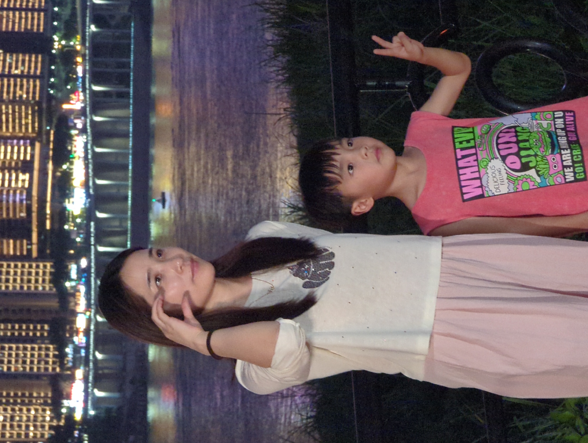 A woman and a child are posing for a photograph at night with a city skyline and a river in the background. The woman, on the left, is wearing a white long-sleeved shirt and a pink skirt. She has her hand near her face and is looking to her left. The child, on the right, is wearing a pink sleeveless shirt with graphics and is holding up two fingers in a peace sign. The background features a brightly lit city with tall buildings and a bridge spanning a river. The area where the subjects are standing appears to be an outdoor grassy area with some vegetation and a railing. The setting is likely the riverfront area of Jiangbeicheng Street, China, given the prominent cityscape.