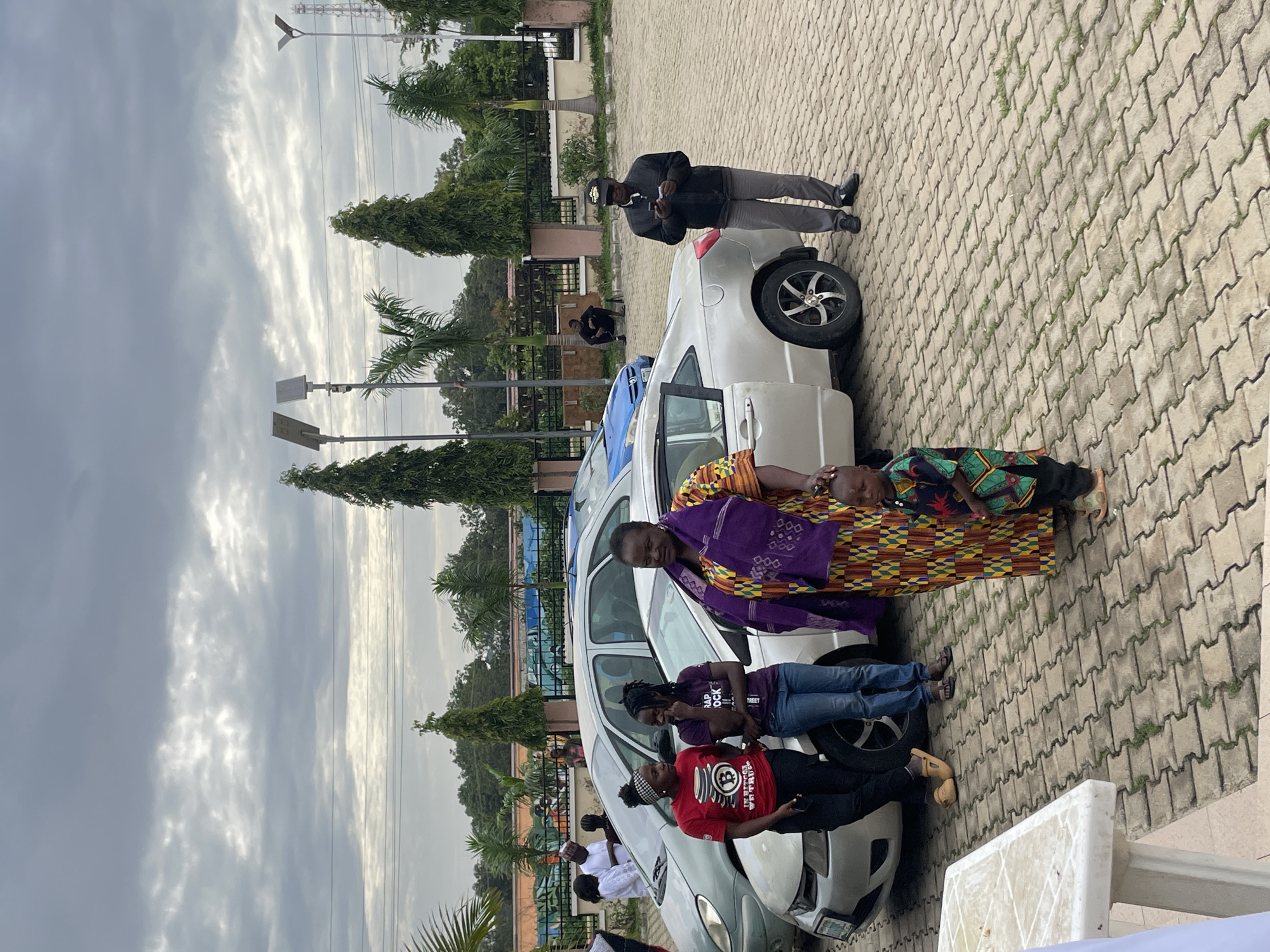 A group of people are gathered around two cars parked on a paved lot. The scene appears to be outdoors, with trees and a building in the background. The sky is cloudy, suggesting it might be overcast or late afternoon.
In the foreground, several people are standing and interacting. A woman in a vibrant, patterned outfit and a purple headscarf is at the center. To her left, two younger women are leaning against a white car, one wearing a purple t-shirt with text, and the other in a red t-shirt with a Bitcoin logo and text. A young child in a colorful garment lies on the hood of the white car, being attended to by an adult. Another man in a black jacket and cap is standing near the rear of the white car, holding something in his hand. Further back, a person in a white garment is visible near another car.
The activity appears to be a social gathering or possibly a community event. The individuals are dressed casually, and their interactions suggest a relaxed atmosphere. The presence of the cars indicates they may have arrived or are preparing to depart. The Bitcoin logo on the red t-shirt is a notable detail. The setting suggests an urban or semi-urban environment in Barakin Lerawi, Nigeria, given the architectural style of the building and the types of trees.