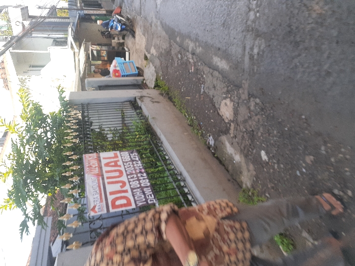 The image shows a person wearing a brown and white patterned shirt and brown pants walking on a narrow street in Cimenyan, Indonesia. The person is walking past a black metal gate with a sign that reads "Dijual" which means "for sale" in English. The sign also includes phone numbers for YUDHA and DEWI. The gate leads to a property with a white painted wall and a small house. There is a green bush in the foreground. The street is paved with asphalt but has a gravel patch. In the background, there are two people standing near a small cart. There is a motorcycle in the background. The street is narrow and appears to be in a residential area. The sky is overcast. The location is in the city of Cimenyan, Indonesia, based on the provided coordinates: -6.888 latitude and 107.652 longitude.