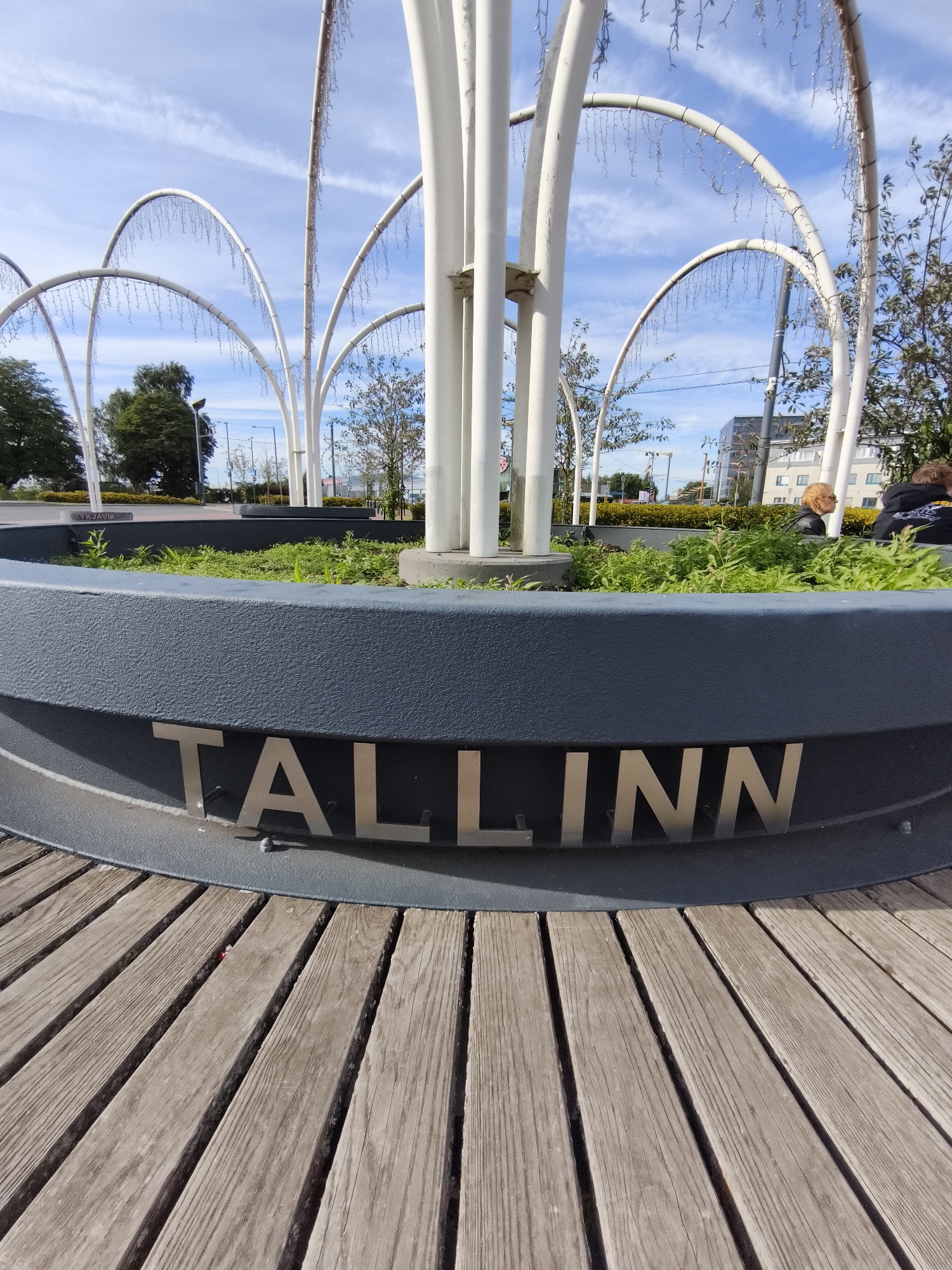 The image depicts a segment of the "Tallinn" lettering, which is mounted on a dark gray, curved wall, likely part of a public art installation or monument. The lettering is in silver, metallic, three-dimensional characters. Below the wall is a wooden deck made of evenly spaced planks running diagonally from the bottom left to the top right of the frame. Behind the wall, a circular planter filled with green vegetation is visible, with tall white vertical columns and arched structures adorned with hanging lights extending upwards. In the background, a clear blue sky with wispy clouds forms the backdrop. Beyond the installation, trees, buildings, and streetlights suggest an urban environment. Two individuals are partially visible in the middle ground, to the right, with one appearing to be a woman with blonde hair and the other wearing a dark hooded sweatshirt. A small red object is visible in the gap between two wooden planks in the foreground. The overall scene is set in Tallinn, Estonia, likely in a public square or park, given the monumental lettering and urban landscaping.