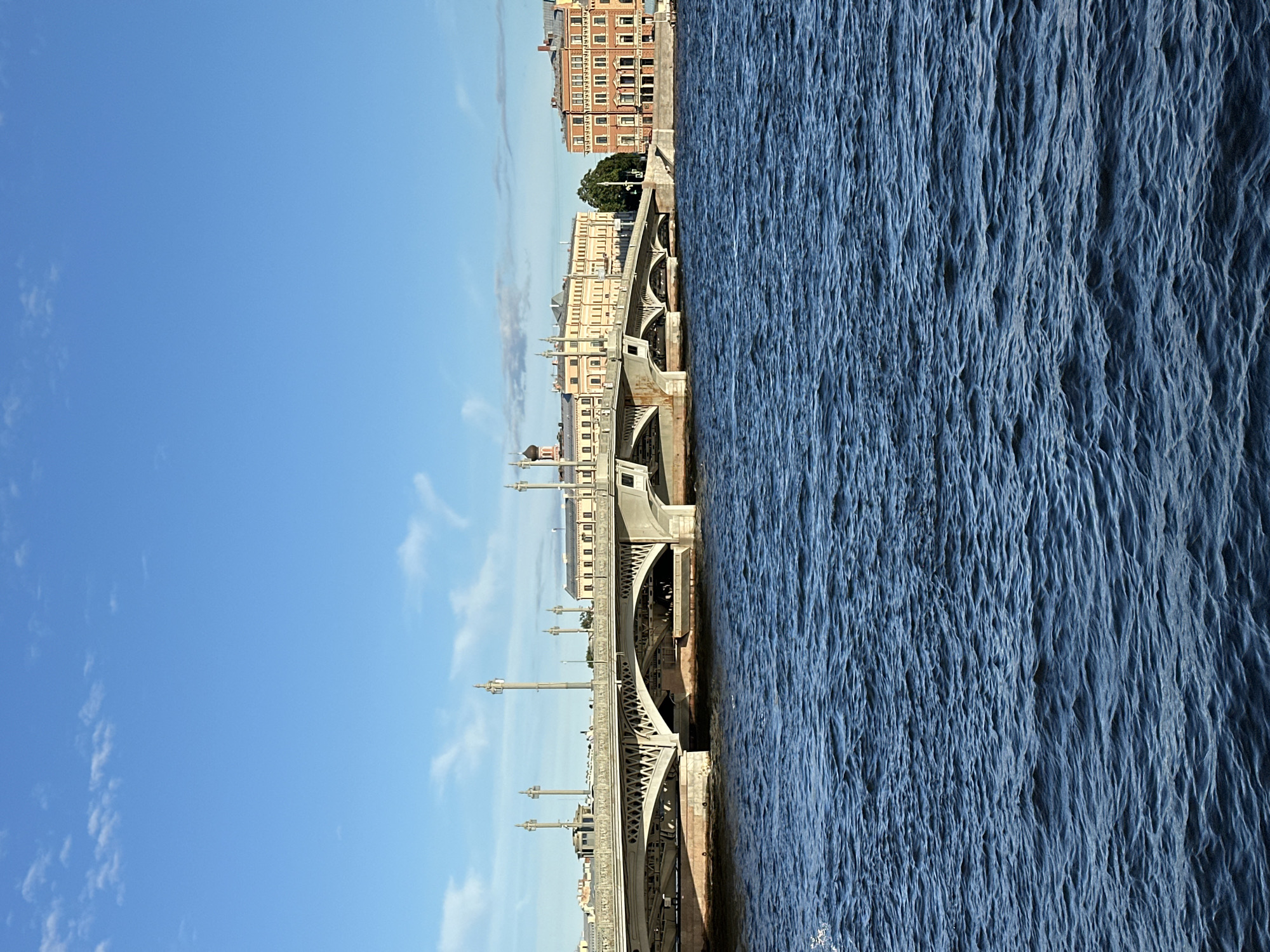 The image captures a daytime view of the Troitsky Bridge spanning the Neva River in Saint Petersburg, Russia. The bridge, made of stone and metal, features multiple arched spans and is lined with ornate lampposts. Buildings with distinct architectural styles line the riverbank behind the bridge. To the left, a long, pale yellow building with many windows and decorative elements dominates the skyline. A dome, possibly part of a church, is visible above this structure. To the right of the bridge, a reddish-brown building with a more structured facade and numerous windows stands prominently. The Neva River in the foreground is a deep blue, with visible ripples indicating a slight breeze. The sky above is a clear, bright blue with scattered wispy white clouds. The scene depicts a calm, urban waterscape under a sunny sky.