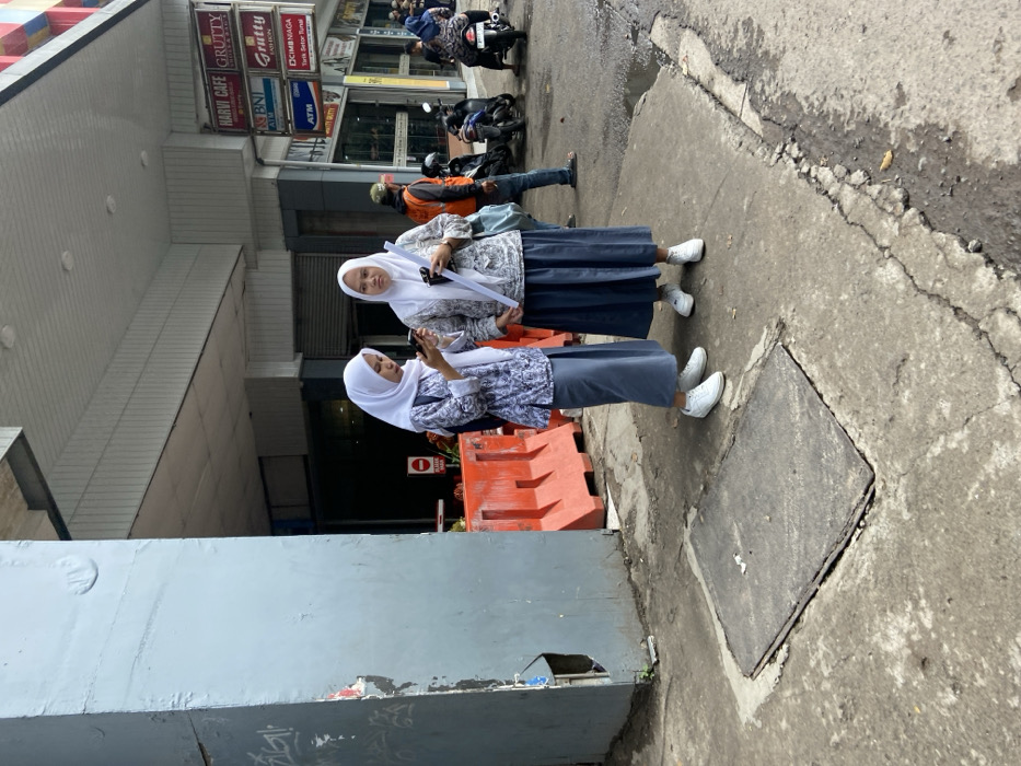 Two women, both wearing white headscarves and long grey skirts, are standing on the sidewalk in front of a building in Bandung, Indonesia. The women are both looking at their phones and are standing next to each other. There are orange construction barriers to their left and a large metal grate in the foreground. There are signs in the background for a cafe, ATM, and a bank. A man wearing a blue shirt and a red cap is walking in the background. There are parked motorcycles behind him. The sidewalk is grey and uneven, and there are cracks running through it. The building in the background is white with a gray roof. There is a sign that reads "Harvi Cafe." The image is taken from a slightly elevated perspective. The location is in the city of Bandung, Indonesia, which is located at the coordinates -6.952, 107.593.