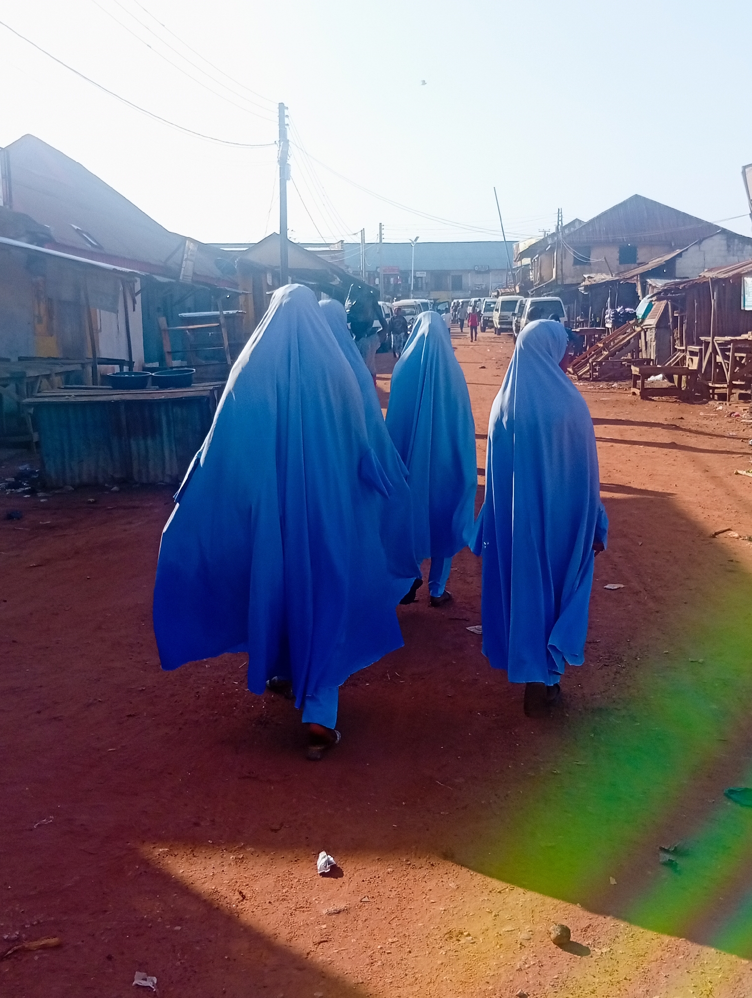 In Bukuru, Nigeria, three individuals wearing bright blue abayas walk away from the camera on a dirt road. The figures are partially obscured by their flowing garments, with only their feet and parts of their legs visible beneath the blue fabric. They are walking in single file down the center of a dusty street lined with market stalls and structures constructed from corrugated metal and wood. The sunlight casts long shadows across the reddish-brown ground, which is littered with small debris. In the background, a row of parked vans and other vehicles suggests a busy thoroughfare, and utility poles with visible wires stretch across the bright, hazy sky. The scene depicts a typical day in a Nigerian market town, with a focus on the draped figures moving through the bustling environment.