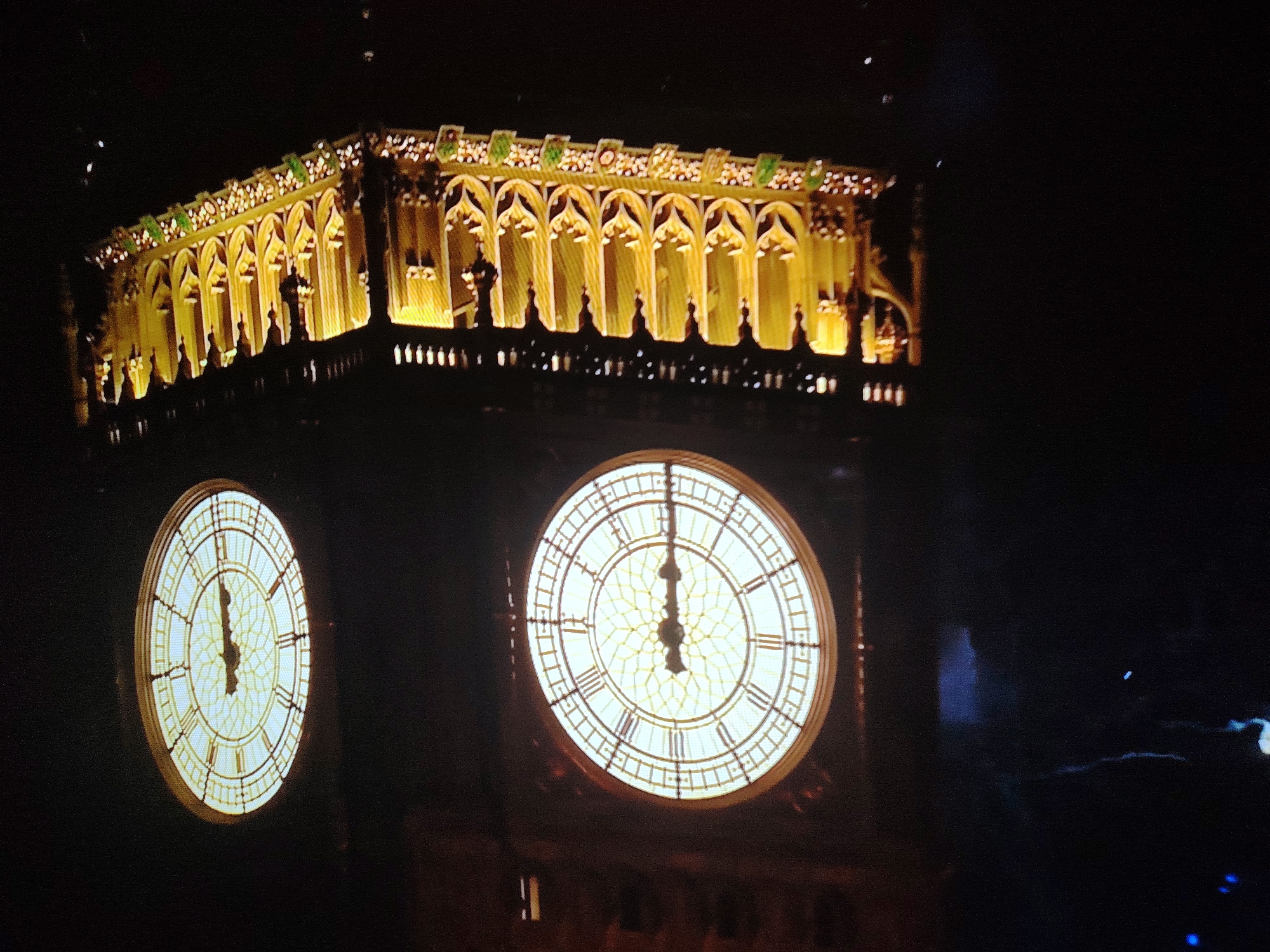 This image captures a night view of the Elizabeth Tower, commonly known as Big Ben, in London, United Kingdom. The iconic clock tower is illuminated, with its large clock faces prominently displayed. The top section of the tower, featuring gothic architectural elements and ornamental details, is also brightly lit with a warm yellow light. One clock face is larger and more centrally positioned in the frame, showing the time as approximately 12:00. A second, smaller clock face is visible to the left, also appearing to indicate 12:00. The background is dark, indicating nighttime, with some faint lights or reflections in the lower right portion of the image. There are no people or active interactions visible in the scene.
