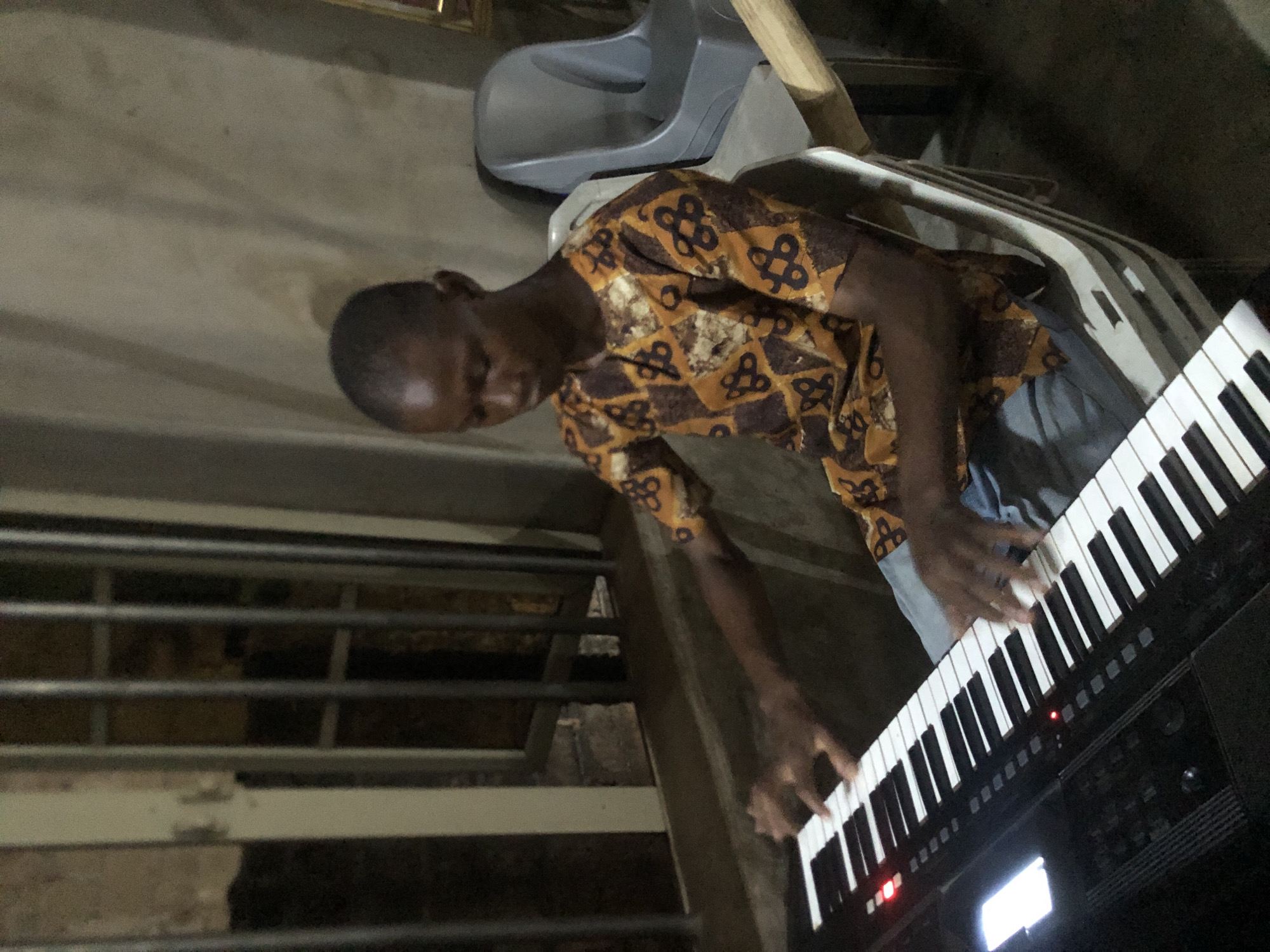 An image, correctly oriented, depicts a young man playing a black digital keyboard in an indoor setting. He is positioned in front of the instrument, with both hands on the keys, seemingly engaged in playing music. The man has dark skin and short hair, and is wearing a short-sleeved shirt featuring a distinctive brown, gold, and black geometric pattern, possibly an Ankara fabric, along with light blue trousers.
The keyboard displays small red indicator lights and a glowing screen, suggesting it is powered on. The overall lighting in the room is dim. In the background, to the left of the man, a structure composed of horizontal metal bars is visible, possibly part of a railing or window grill. Behind him, a grey plastic chair is partially visible, along with other indistinct elements of what appears to be a utilitarian or minimally furnished room. The man's expression is one of focus on the activity. No discernible text is visible within the image. The scene is likely set in Ukwu-Abwa, Nigeria.