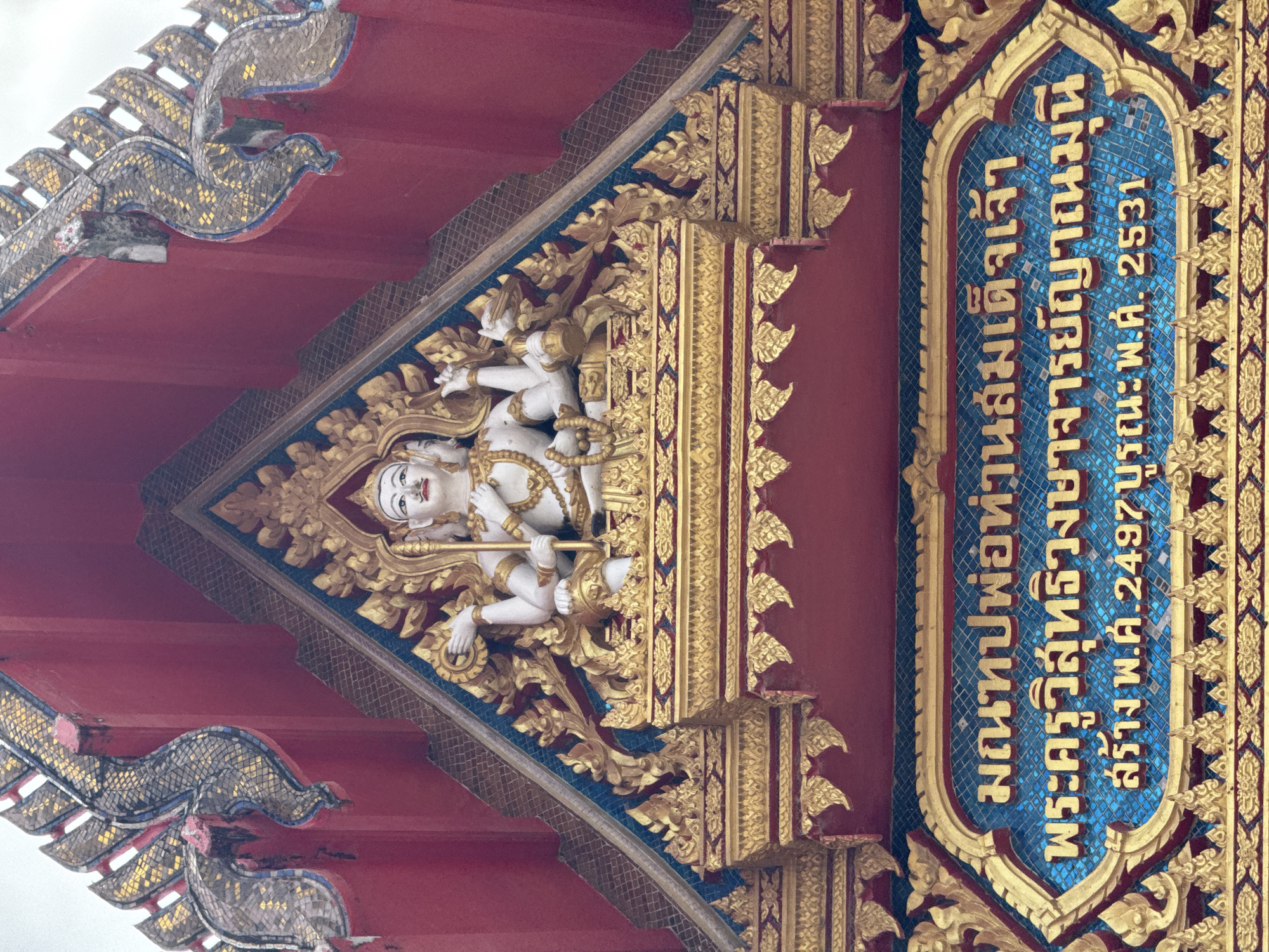 The image shows the upper facade of a temple in Chalong, Thailand. A gilded, multi-armed statue of a deity is prominently featured at the apex of the structure, adorned with intricate gold ornamentation. Below the statue, blue tiles form a backdrop for Thai script inscribed within a gold border. The script, transliterated, reads: "Manthop Pho Than Somdet Chao Phra Khru Wisutthi Rangsithan Muni." Below this, it states: "Khrang, Ph.S. 2497, B.E. 2531." The architecture displays vibrant red walls and ornate, serpent-like decorative elements, also covered in gold and mosaic tiles. The overall scene is a detailed view of religious architecture and statuary.
