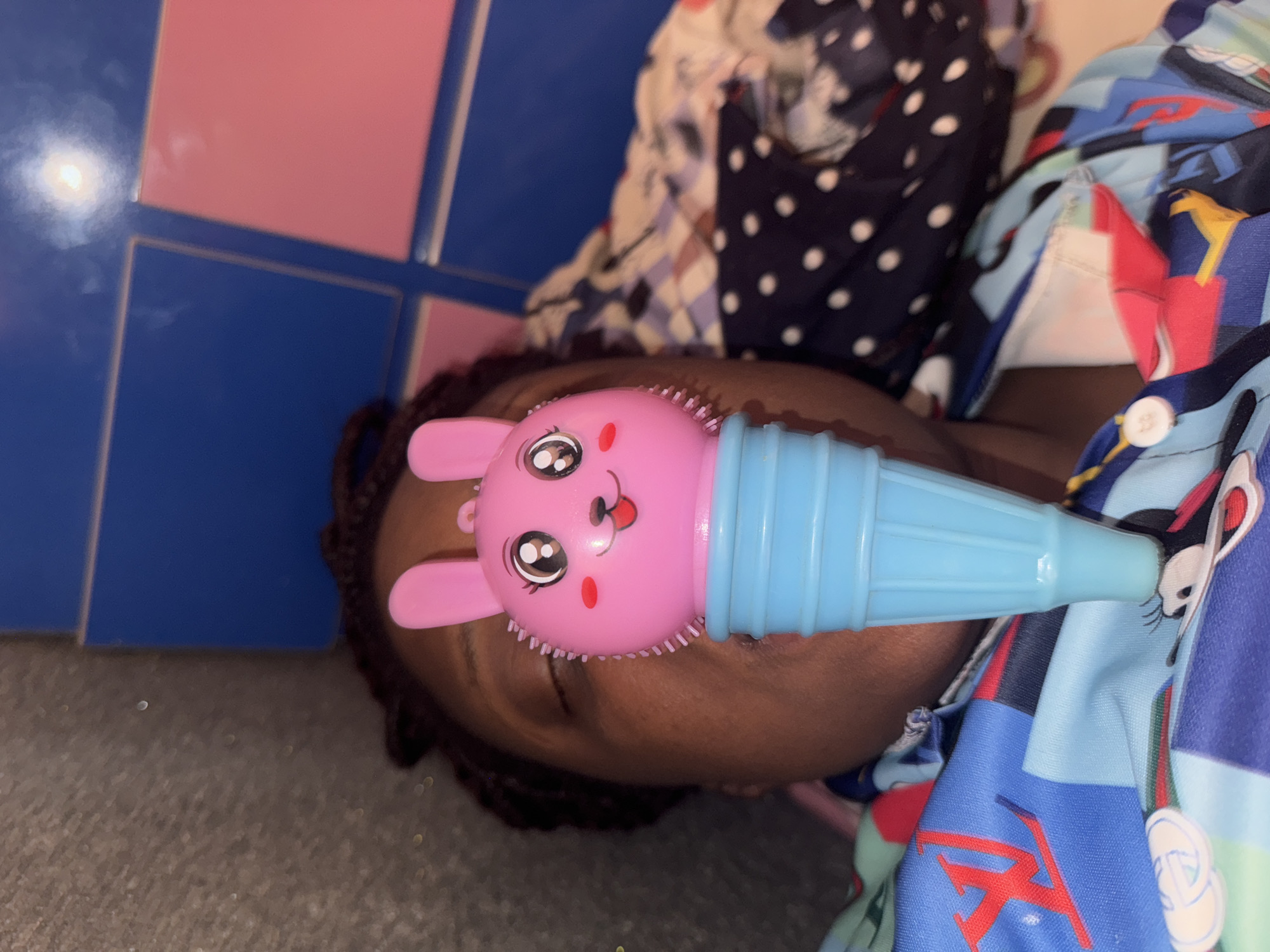 A close-up, tilted shot reveals a child lying down, with a pink and blue rabbit-shaped object positioned near their face. The child's hair is dark and braided. The child is wearing a patterned shirt with various colors and designs, including blue, red, and white elements. Part of another patterned fabric, predominantly dark blue with white polka dots, is also visible in the background. The floor is mostly gray and reflective, with blue and pink square tiles visible along one edge of the frame. The location, Sabon Tasha, Nigeria, is not evident in the image.