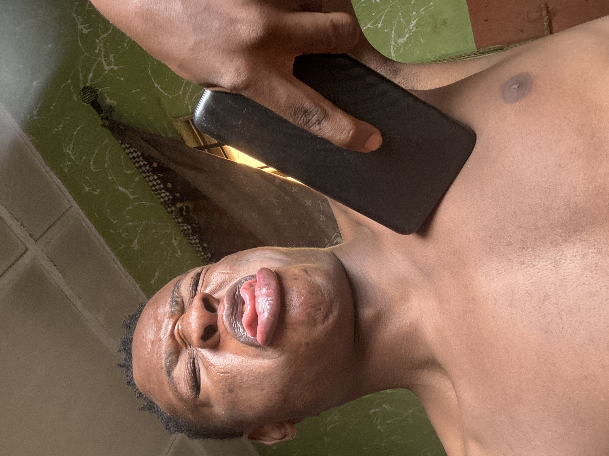 A low-angle, close-up shot shows a Black person with closed eyes and a contorted facial expression, their tongue sticking out. A dark-colored object, likely a smartphone, is held against their chest by another person's hand. The background features a green wall with white streaks and a portion of a patterned curtain. The ceiling, visible at the top, has a grid pattern. The scene appears to be indoors, possibly in a home in Ikorodu, Nigeria.