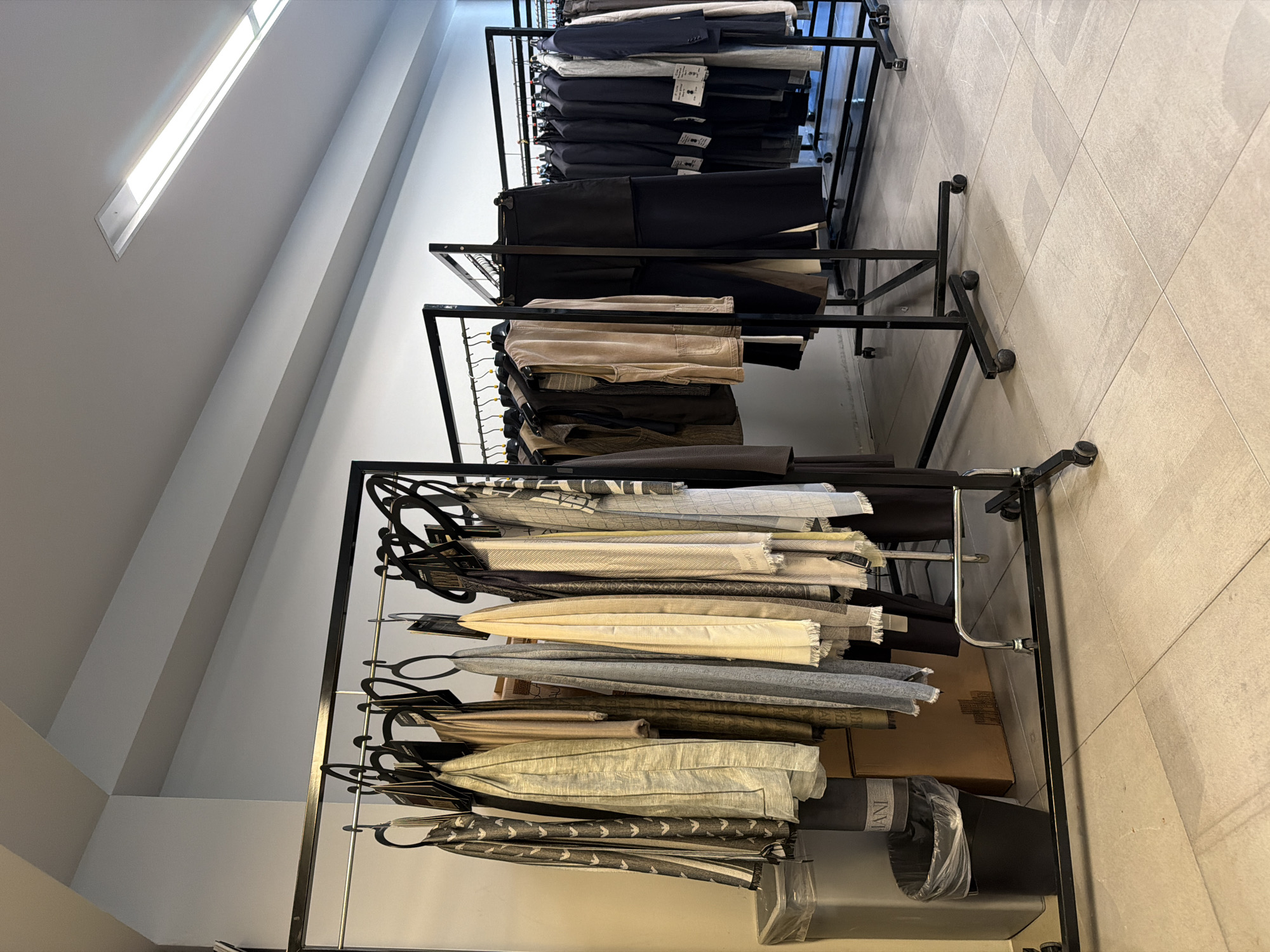 The image depicts an interior scene, likely a clothing store or showroom, in Milan, Italy. Multiple black metal clothing racks are set up on a light-colored tiled floor. The racks are laden with various garments, primarily scarves and formal wear such as suits and jackets. The color palette is subdued, dominated by neutral tones like grey, beige, and black, with some items featuring subtle patterns. In the foreground, on the left side, a tall silver waste bin and a smaller black trash can are visible. Behind the clothing racks, a light grey wall serves as the backdrop, with a long fluorescent light fixture visible on the ceiling. The overall impression is one of an organized retail or display space, emphasizing a sophisticated and minimalist aesthetic.