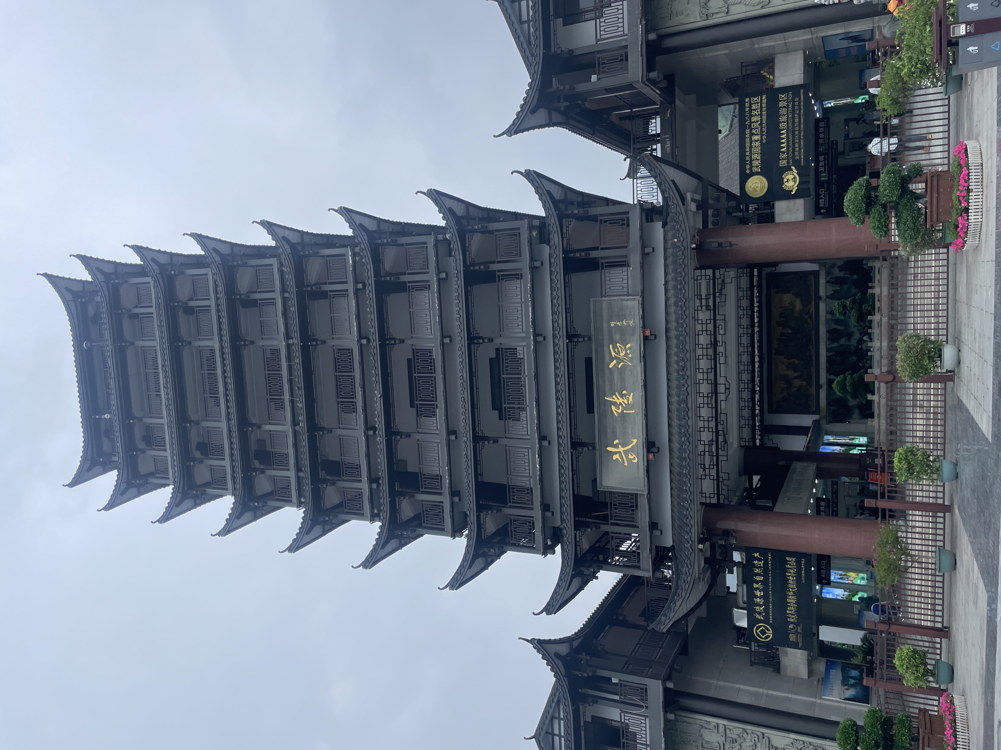 The image shows a tall, traditional Chinese building in Wulingyuan, China. The building has multiple levels, each with a curved roof. There is a sign with Chinese characters on it. To the right of the building, there is a sign indicating that it is a National AAAAA tourist attraction and also indicating the Wulingyuan World Natural Heritage site. Additional signs advertise that it is a key scenic area. The sky is overcast. A sign indicates entrance for line A, as well as information about toilets and boiled water. There are small potted plants surrounding the base of the building.