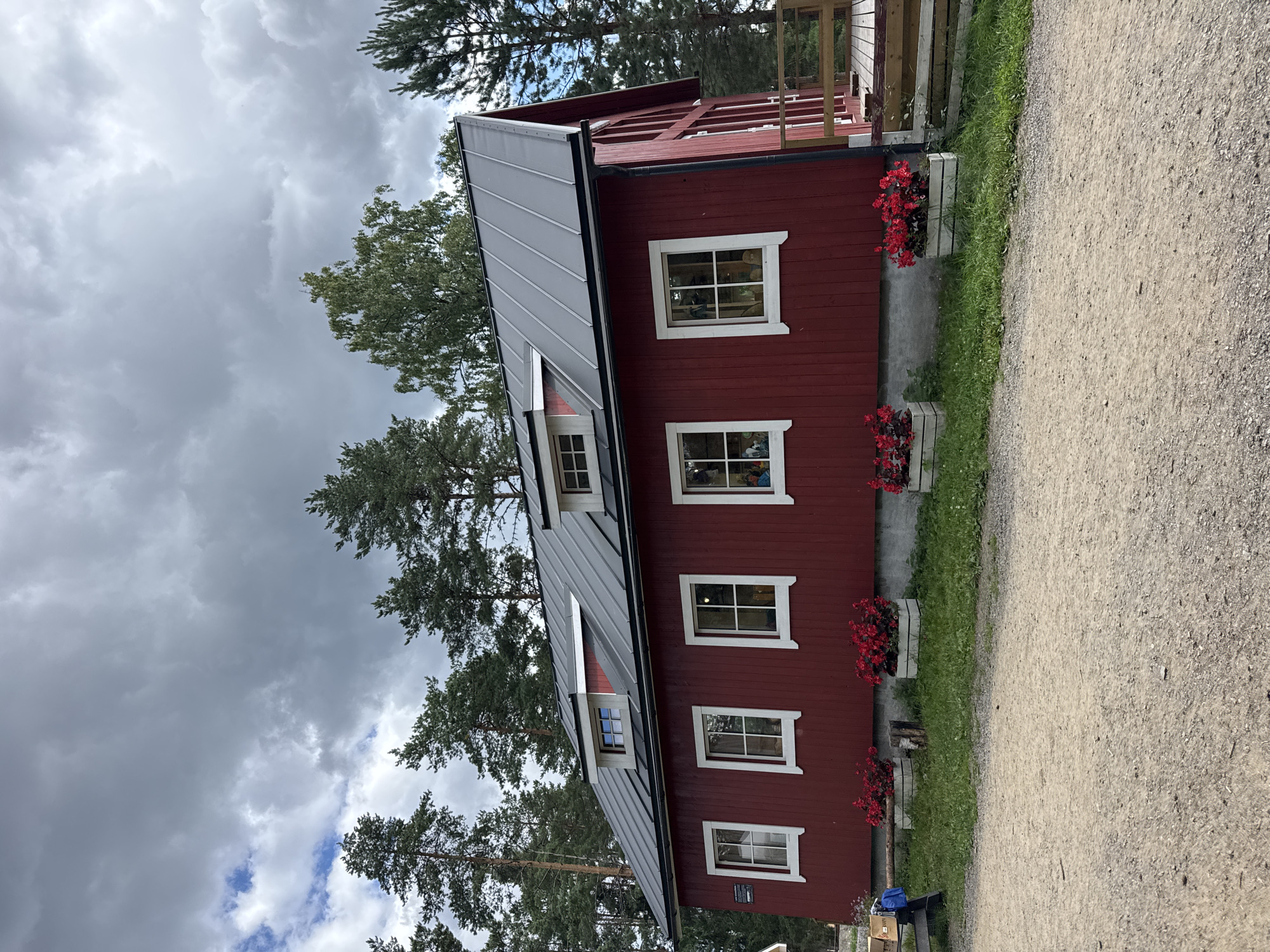 In the city of Khankanmyaki, Russia, a bright red, wooden, two-story building stands under a cloudy sky. The building features a dark gray, sloped roof with multiple dormer windows. The side facing the viewer has five white-framed windows on the first floor. In front of these windows are planters filled with vibrant red flowers. A gravel pathway runs along the front of the building, and to the left, a person wearing a blue backpack is visible. The building is surrounded by tall pine trees. To the right, a wooden deck with railings extends from the building.