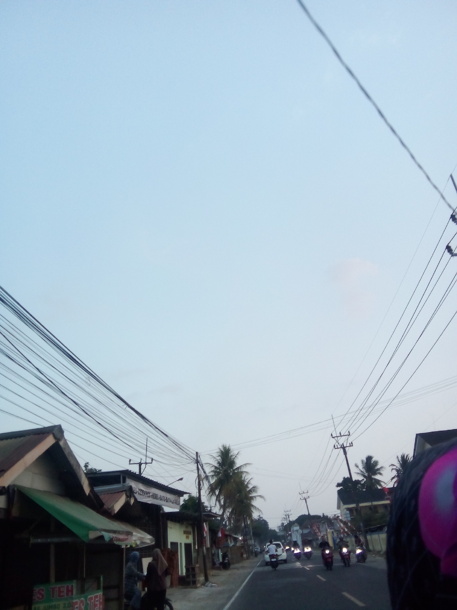 This image depicts a street scene in Carita, Indonesia, likely during dusk or early morning. The sky is a pale blue, with power lines crisscrossing overhead. On the left side of the road, there are several small buildings, one with a green awning and signage indicating "ES TEH". People are visible on the sidewalk, some walking and one riding a motorcycle. The road itself has traffic, with several motorcycles and a white car. The environment appears to be a mix of residential and commercial buildings, with palm trees scattered throughout. The foreground is partially obscured by what appears to be the helmet of the person taking the photograph.