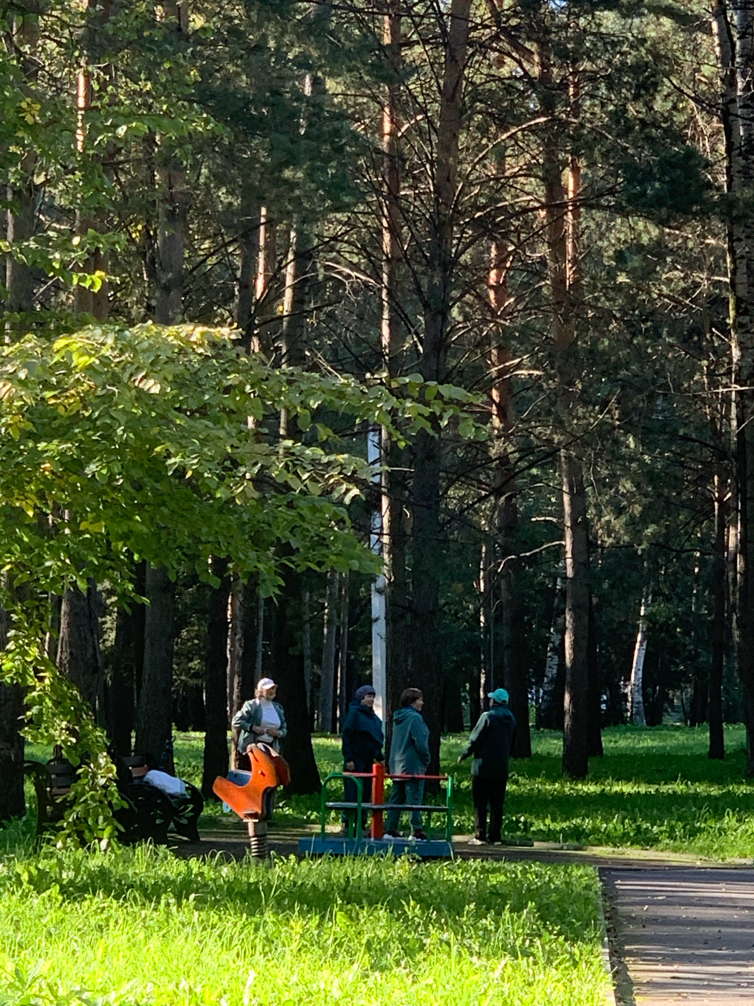 The image depicts a sunny park scene with dense coniferous trees in the background, characteristic of the region. In the foreground, a paved path borders a grassy area. Several people are present: a woman in a white cap and green jacket stands near a bright orange spring rider on a playground. To her right, two individuals in hooded jackets stand near a merry-go-round. To the left, a person sits on a park bench partially obscured by foliage. The overall atmosphere is casual and recreational, with the park appearing well-maintained. Based on the presence of specific playground equipment and the general ambiance, the location is likely a public park or recreational area within the city of Prokopyevsk, Russia, possibly in a residential neighborhood or a dedicated green space.