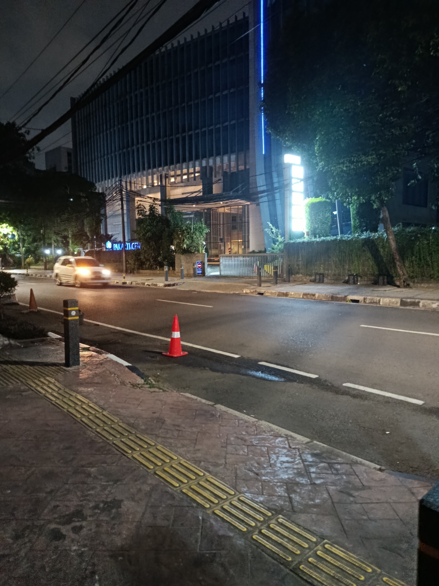 The image shows a nighttime street scene in Central Jakarta, Indonesia, likely on Jalan Professor Doktor Satrio. On the right side of the image, a modern building with a facade of vertical louvers is illuminated by a blue light strip and a bright white sign. The street is wet, reflecting the ambient light. A white SUV is seen driving on the road, its headlights on. Orange traffic cones are placed on the side of the road and on the sidewalk. The sidewalk features tactile paving for the visually impaired. Trees and dense greenery flank the building and line parts of the street. The sky is dark, indicating it is nighttime.