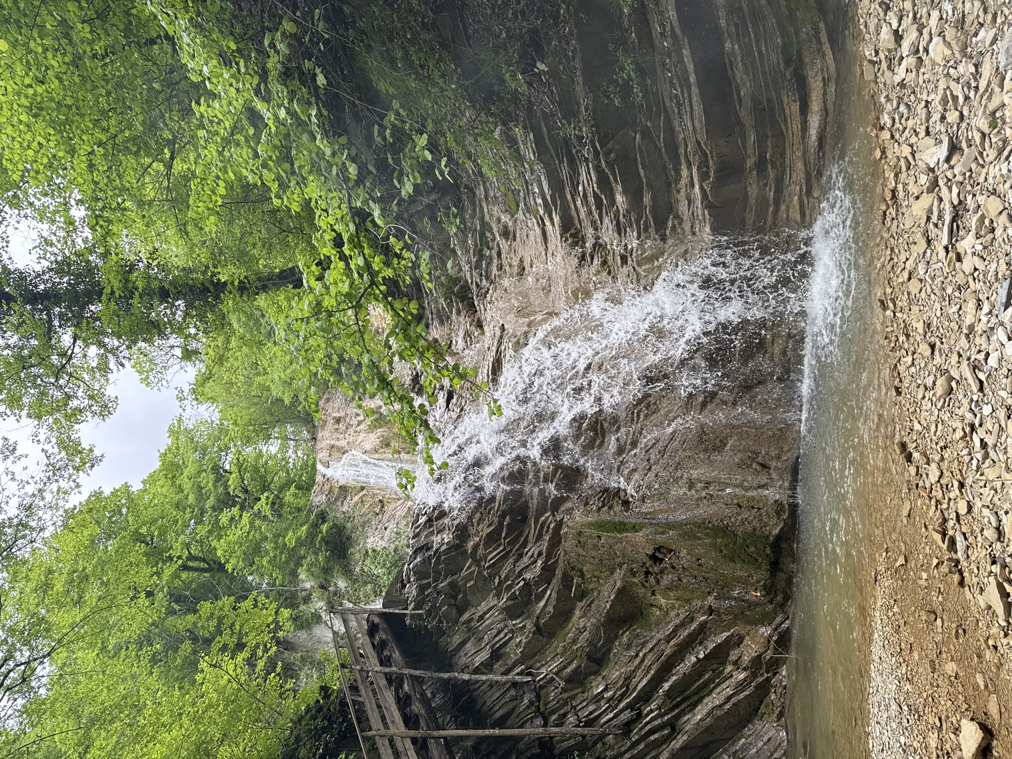 The image depicts a waterfall cascading over a rocky cliff face in a natural environment, likely located in the vicinity of Podkhrebtovoe, Russia. The cliff is composed of layered sedimentary rock. Lush green foliage dominates the left side of the frame. A crude wooden ladder-like structure is visible near the base of the cliff. The waterfall's water impact creates a splash zone, and a pool forms at the base of the falls. The right side of the frame shows a gravelly bank adjacent to the water. There are no people visible in the image. The overall scene suggests a natural, potentially remote location.