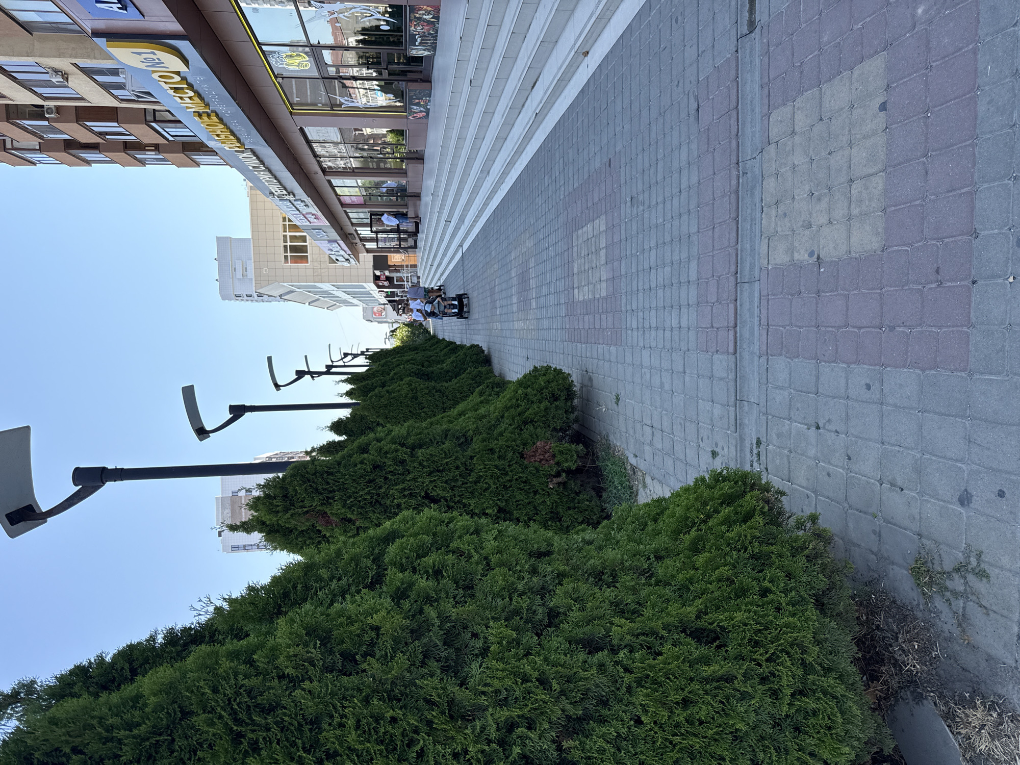 In Anapa, Russia, this image captures a sidewalk lined with cypress trees on the left and a modern building with shops on the right. The ground is paved with square interlocking bricks in shades of grey and reddish-brown, forming a pattern. A wide staircase leads up to the building's entrance, which has large glass windows and doors. Several people are visible on the sidewalk and stairs. Two individuals are walking together, with one pushing a stroller that contains a person. Another person stands near the stairs. Overhead, contemporary streetlights with solar panels are mounted on tall poles. The sky is clear and blue, indicating a sunny day. The building features signs with Russian text, one of which reads "РЫБНОЕ МЕСТО №1" which translates to "FISH PLACE NO. 1". The scene appears to be a commercial or pedestrian area within the city.