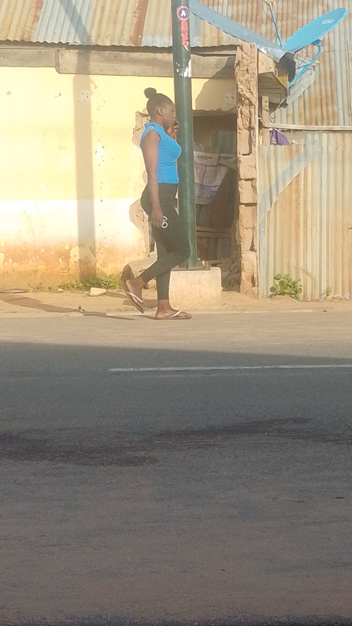 In a medium shot, a young Black woman walks along a paved road in Tempure, Nigeria. She is wearing a bright blue sleeveless top and dark leggings with white accents. Her hair is pulled up in a bun. She is walking from left to right across the frame, and her body is angled slightly away from the camera. She wears white flip-flops.
The setting appears to be an urban or semi-urban area. Behind the woman, there is a building with a light yellow facade and a corrugated metal roof. Part of the building is constructed with rough stone. A black metal pole with some graffiti stands in front of the building, and a satellite dish is mounted on the roof. The ground in front of the building is dirt with some sparse vegetation. The road in the foreground is asphalt and shows signs of wear. The lighting suggests it is daytime, likely morning or afternoon, with distinct shadows cast by the building and the woman. The overall mood is casual and everyday. There is some text visible on the woman's leggings, appearing as two concentric circles with white outlines. There is also a faint "A" logo visible on the pole.