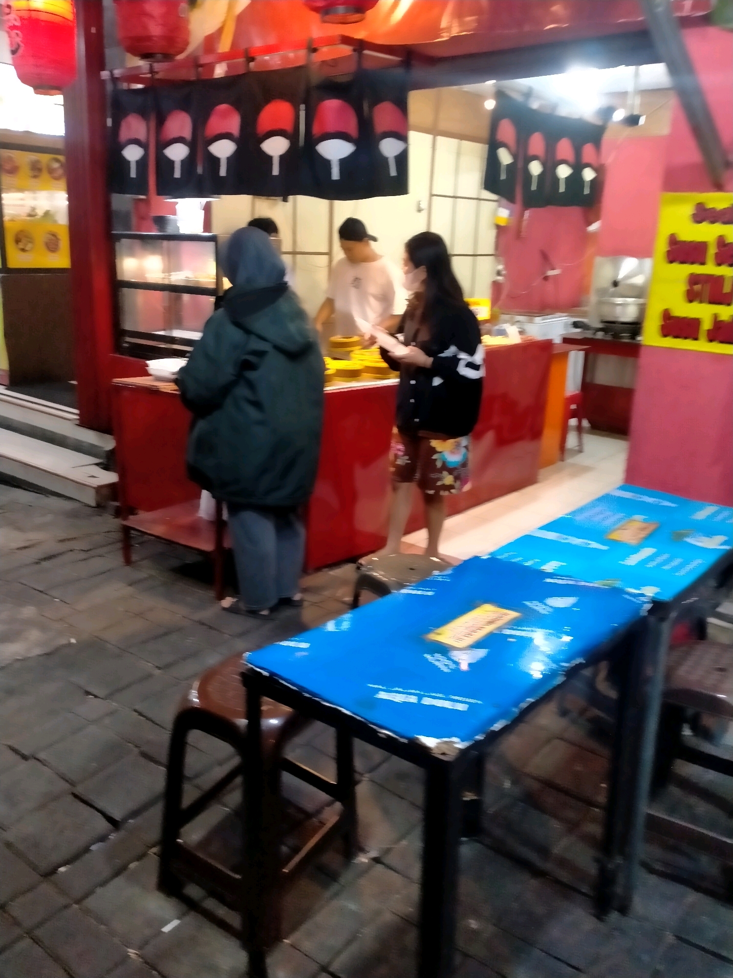 This image depicts an outdoor food stall at night, possibly located in Batu, Indonesia, as indicated by a yellow sign in the upper right with text that may be in Indonesian. The stall features red banners with a black and white symbol, potentially related to anime culture, hanging above a display case. Two individuals are visible at the stall; one wearing a dark green hooded jacket and grey pants, and another wearing a black and white patterned sweater and floral shorts. A third person, wearing a white t-shirt and a black cap, is also partially visible behind the display case. In the foreground, there are two blue-covered tables with chairs, one of which is a dark brown stool. The ground is paved with interlocking stones. The overall scene suggests a casual dining atmosphere.
