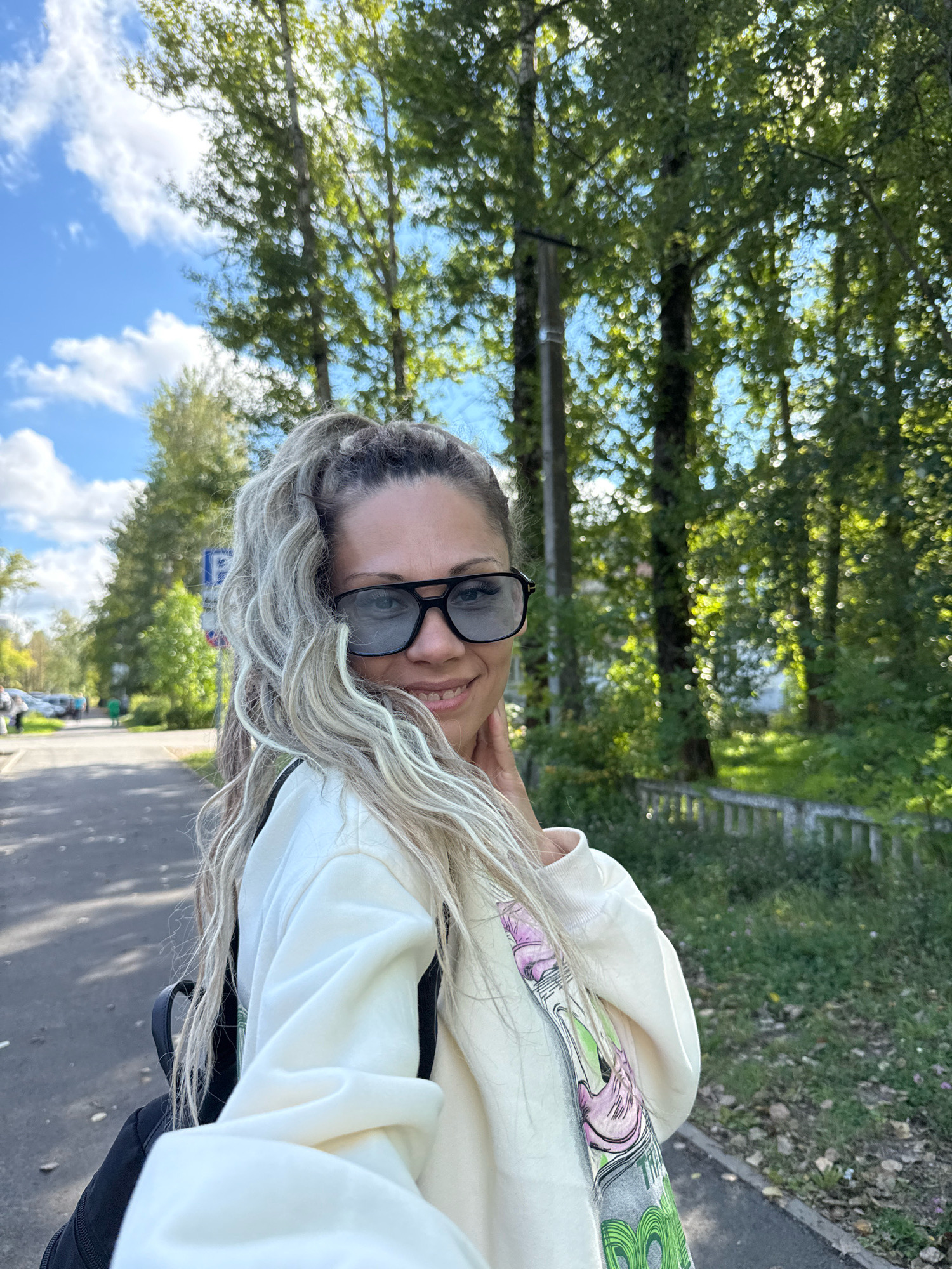 A woman with blonde dreadlocks pulled into a ponytail, wearing black sunglasses and a cream-colored sweatshirt with a graphic print, smiles for a selfie outdoors. She is standing on a paved path, possibly a sidewalk, with trees and grass to her right and a road with cars and a blue parking sign to her left. The sky is blue with scattered white clouds. The location is identified as Rusanovo, Russia.
