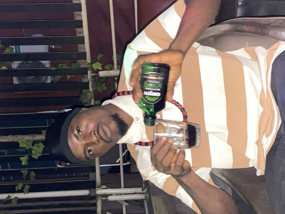A man in a striped shirt pours a bottle of “Origin” alcohol into a glass. He wears a black head covering and a necklace made of small red and black beads. The background features a partially visible structure with horizontal slats and artificial ivy. The lighting suggests an indoor or nighttime setting.