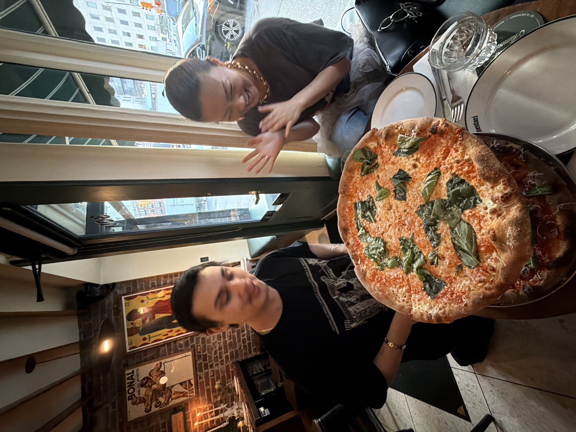 The scene is an indoor dining setting in a restaurant, likely in New York City, United States. Two people are present; a woman is sitting near a window, smiling and clapping her hands. She is wearing a dark shirt and a gold chain. A man is seated opposite her, holding a large pizza.
The pizza is topped with tomato sauce, mozzarella cheese, and fresh basil. Next to the pizza are plates, silverware, and a glass of water. A handbag is visible near the woman. Outside the window are city buildings and cars. The background of the man shows interior restaurant decor including brick walls and framed posters.