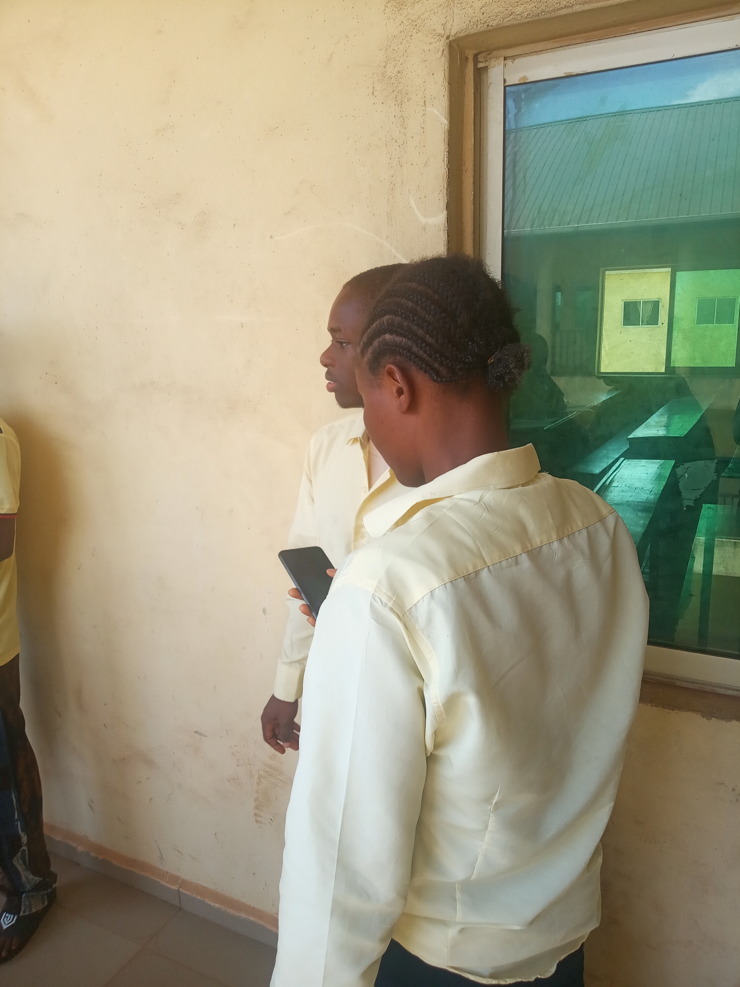 Two individuals, possibly men, are captured standing next to a cream-colored wall and a tinted window, likely in an indoor or covered outdoor setting. The person in the foreground is viewed from behind, wearing a light yellow long-sleeved collared shirt. Their dark hair is styled in braids pulled back. They are holding a black smartphone in their right hand.
To the left, a second person is partially visible in profile, also wearing a similar light yellow long-sleeved shirt. A third person's lower leg and foot, wearing dark sandals, are seen on the far left edge of the frame.
The window features green and blue tinted glass, reflecting what appears to be other nearby buildings with yellow and green exteriors and visible windows, along with a glimpse of a light blue sky or structure. The wall behind the individuals is a plain, light cream color, showing some signs of wear and dirt. A light-colored tiled floor is visible at the bottom.
The scene suggests a casual daytime gathering or waiting period in Nsugbe, Nigeria, with the individuals appearing neutral in mood, possibly conversing or awaiting an event. No text is visible in the image.
