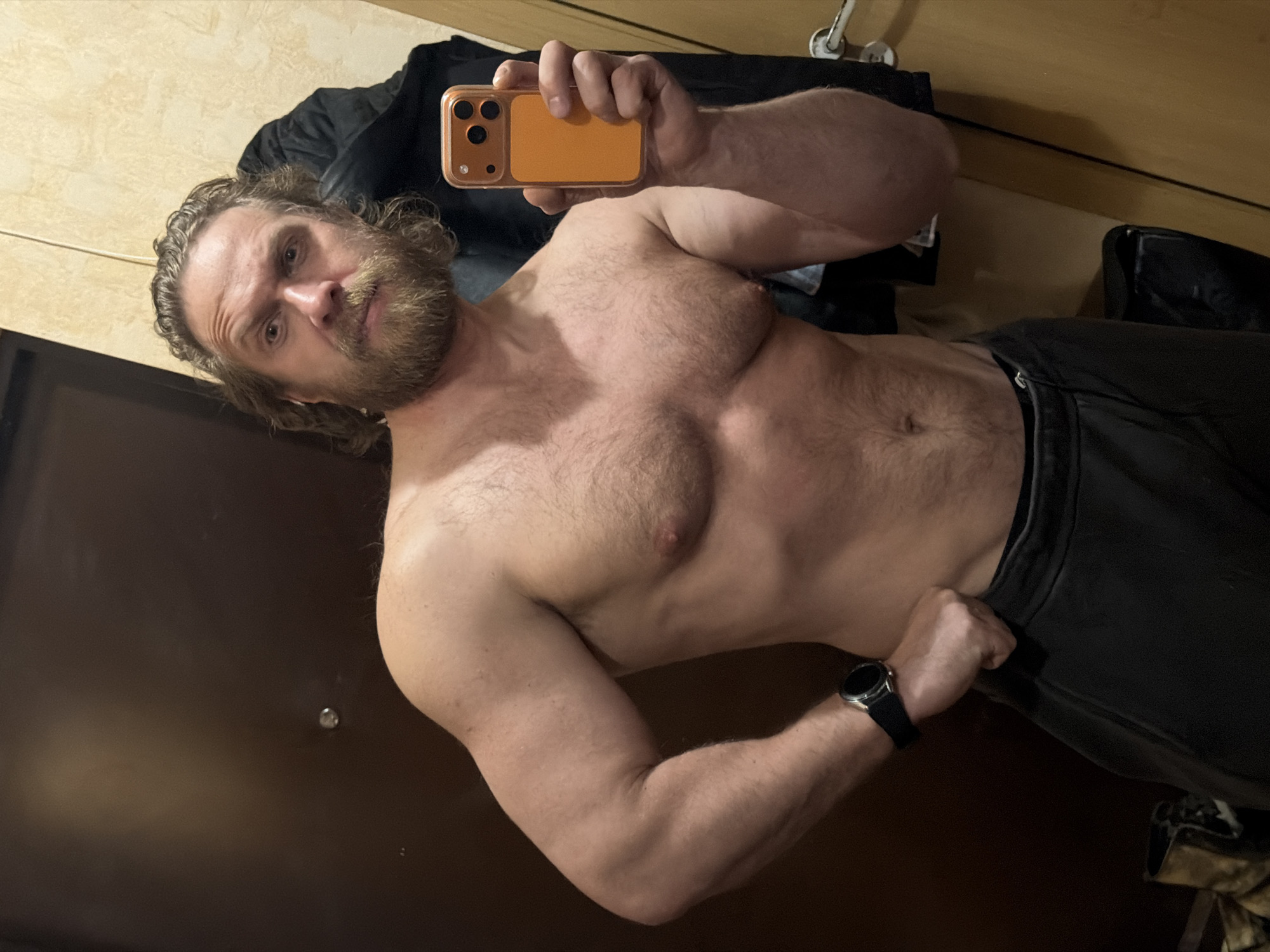 A muscular man with a beard and long hair is taking a selfie. He is shirtless and wearing dark-colored pants. He is holding an orange smartphone. He is also wearing a black smartwatch on his left wrist. The background is a blurred interior with a door on the left and clothing on the right. It is not possible to identify the exact location within Saint Petersburg, Russia.