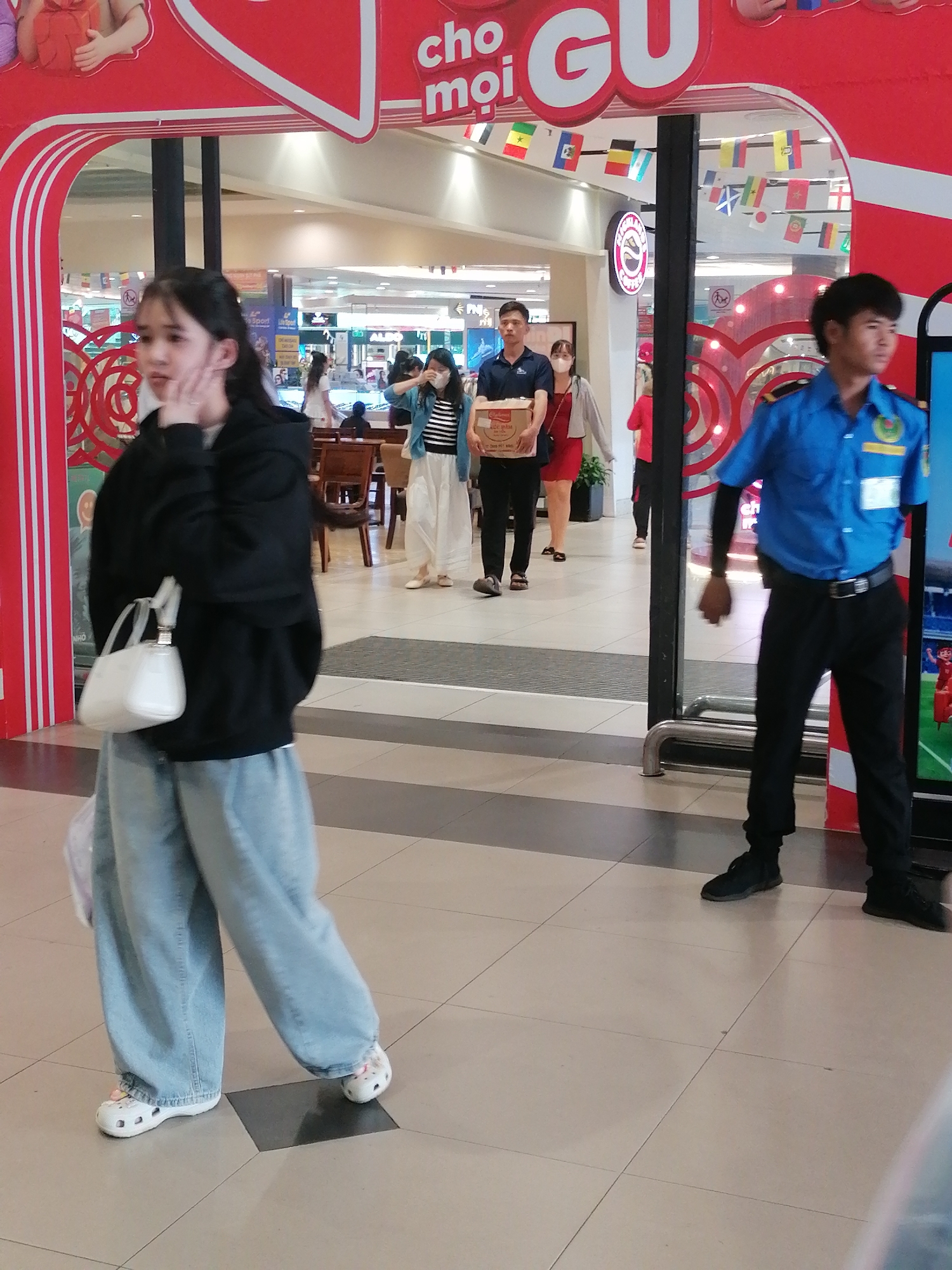 A young East Asian woman with long dark hair walks left to right across a tiled indoor walkway in what appears to be a shopping mall. She is wearing a black hooded sweatshirt, loose light blue jeans, and white slip-on shoes. She is carrying a white handbag on her left shoulder and a clear plastic bag in her left hand. Her right hand is near her face.
To her right, further in the background, a man in a blue uniform with black pants and a black belt walks right to left. He has short dark hair and is wearing black shoes. He appears to be a security guard.
Behind the woman, a man in a dark blue t-shirt and black pants is walking right to left, carrying a cardboard box. A woman in a red dress and a light gray cardigan walks behind him.
The background shows a red and white curved awning with text in what appears to be Vietnamese. Flags are hung across the top of the awning. The floor is tiled with light beige and darker beige square tiles, arranged in a diamond pattern. The lighting is bright and diffused.