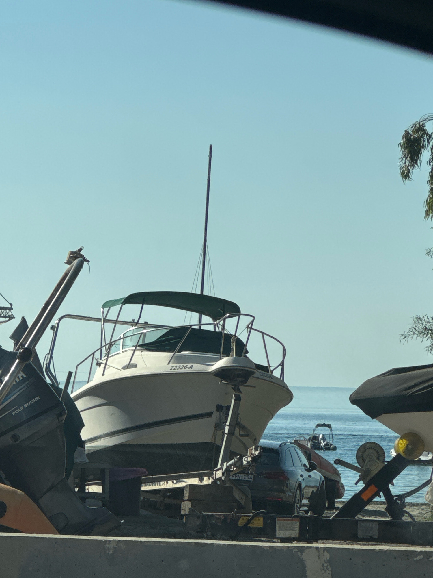 The image captures a sunny day scene at a waterfront location, possibly a marina or boat storage area, with the Limassol coastline in the background. In the foreground, several boats are parked on trailers, some partially covered. A white motorboat with a green canopy is prominently positioned on a trailer. To its left, a portion of another boat with an outboard motor is visible. A dark-colored SUV is parked behind the white motorboat, also on a trailer. In the background, the calm, blue water of the sea extends to the horizon, with a small boat in the distance. Trees with green foliage are visible on the right side of the image, suggesting a coastal environment. The overall impression is of a recreational area by the sea, with boats ready for use or undergoing maintenance.