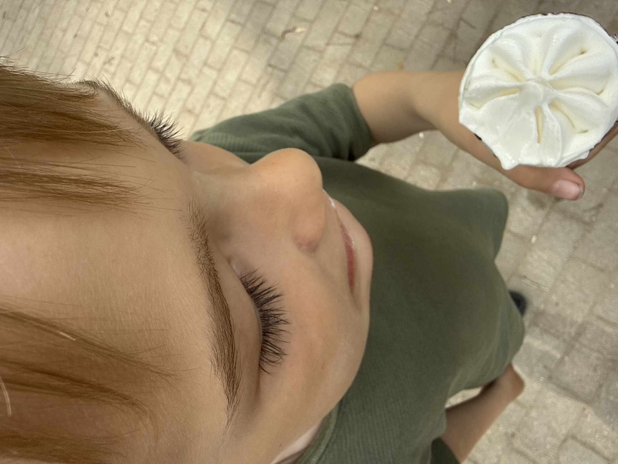 A child with fair skin and light brown hair is holding a cupcake with white frosting. The child is wearing an olive-green shirt. The background shows a light-colored paved surface, possibly a sidewalk. The photograph is taken from a high angle, looking down at the child. The location is unknown.