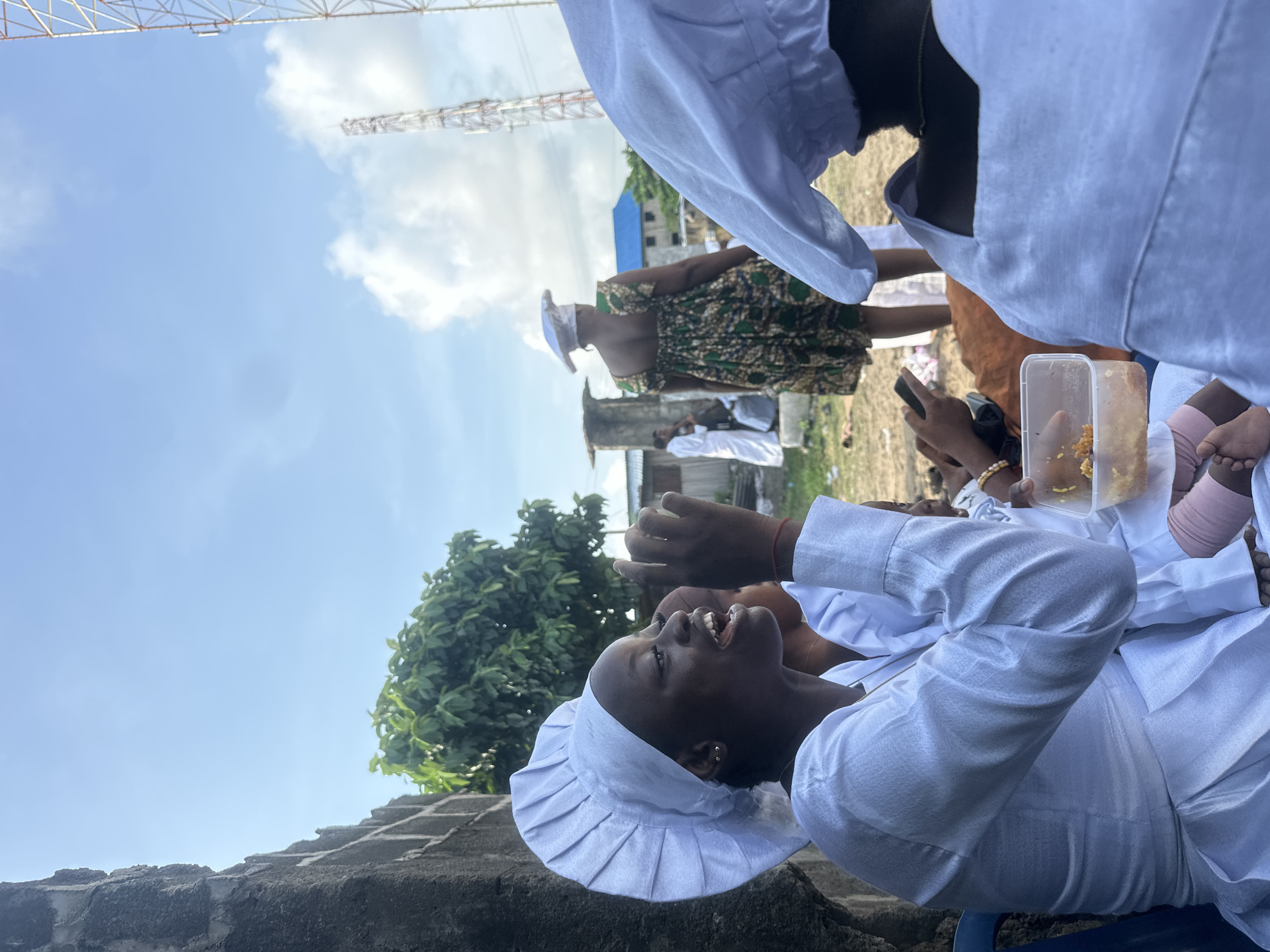 The image depicts an outdoor gathering in Badagry, Nigeria, under a bright, partly cloudy sky.
In the left foreground, a young woman with dark skin is prominently featured from the chest up, positioned against a gray concrete block wall. She wears a white, pleated, chef-style hat and a long-sleeved white garment. Her mouth is open, indicating she is speaking or singing, and her left hand is raised to her mouth, while her right hand is partially visible further back. A small gold earring is visible on her left earlobe. Behind her, another individual in similar white attire is partially visible.
In the immediate lower foreground, parts of two bare feet, one with a pink bandage or fabric wrapping, are visible next to a clear plastic container holding a yellow-orange granular food, likely rice. Hands are partially visible holding both the container and a dark-colored mobile phone.
In the mid-ground, a woman with her back to the camera stands wearing a sleeveless, patterned dress in shades of green, brown, and black, along with a white hat similar to the foreground subject's. To her left, another person in a long white robe is partially visible near a weathered concrete structure. The ground in the mid-ground is unpaved, appearing as dirt or sparse grass.
The background features several structures. To the far left, a dense green tree provides foliage. Towards the center-right, two communication towers, one larger and more prominent than the other, extend into the sky. Scattered buildings with light-colored walls and one with a blue corrugated roof are visible between the figures and the towers. The overall scene is brightly lit by natural sunlight, casting minimal shadows.