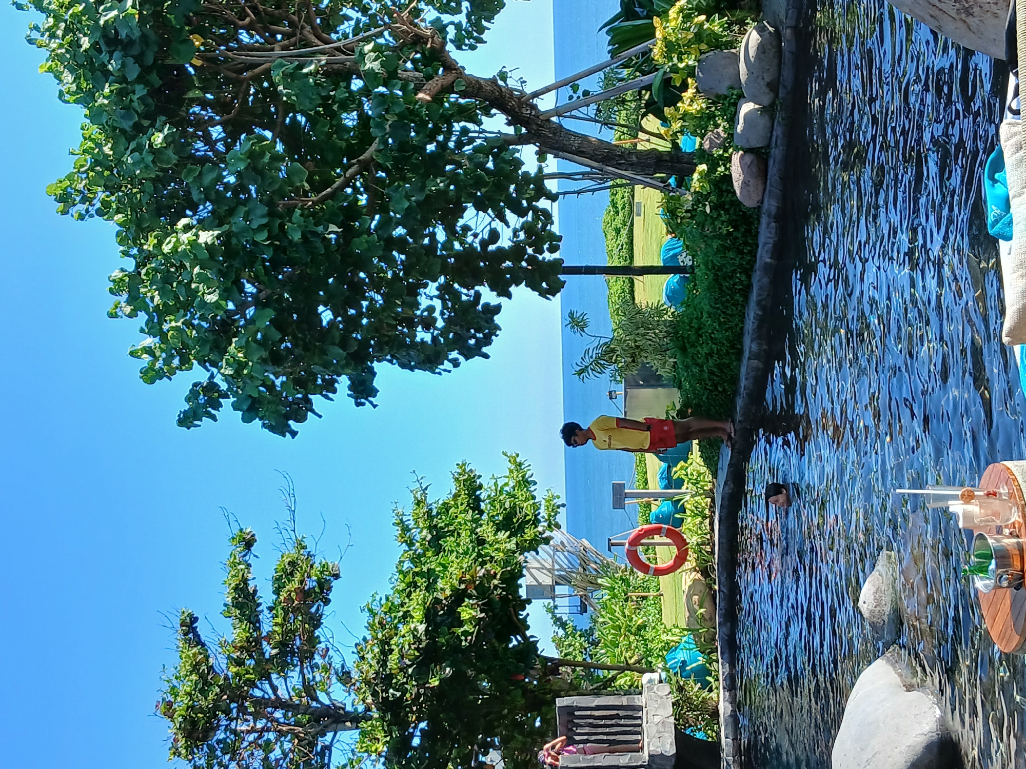 The image depicts a resort swimming pool area in Mengwi, Indonesia, with the ocean visible in the background. In the foreground, a person is swimming in the pool, and a table with drinks and ice bucket is placed near the edge. On the pool deck, a person in a yellow shirt and red shorts, possibly a lifeguard, stands near a red life preserver. Lush green trees and landscaping surround the pool. The scene is bathed in bright sunlight under a clear blue sky.