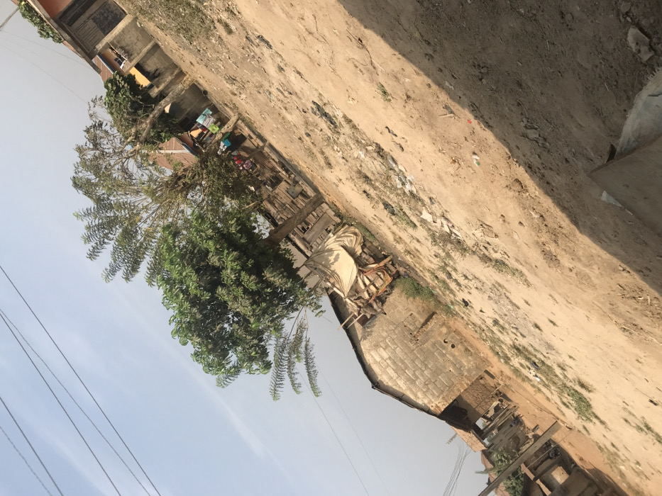 The scene depicts an outdoor, semi-developed area in Ago-Abo, Nigeria, under a clear, hazy light blue sky with power lines visible in the upper left quadrant. The ground consists of uneven, light brown dirt with sparse patches of green vegetation and scattered debris. In the mid-ground left, a structure constructed from grey concrete blocks is visible, adjacent to a makeshift stall or storage area covered with a light-colored fabric tarp and supported by wooden frames. A prominent green-leafed tree stands centrally, with additional trees, including one with elongated leaves, further back. In the background right, residential structures are present, some with unpainted block walls and others with sections of reddish-brown coloring. Laundry is hanging on lines near these structures, and two to three indistinct figures are discernible in the far background. A diagonal shadow extends across the lower right portion of the ground.